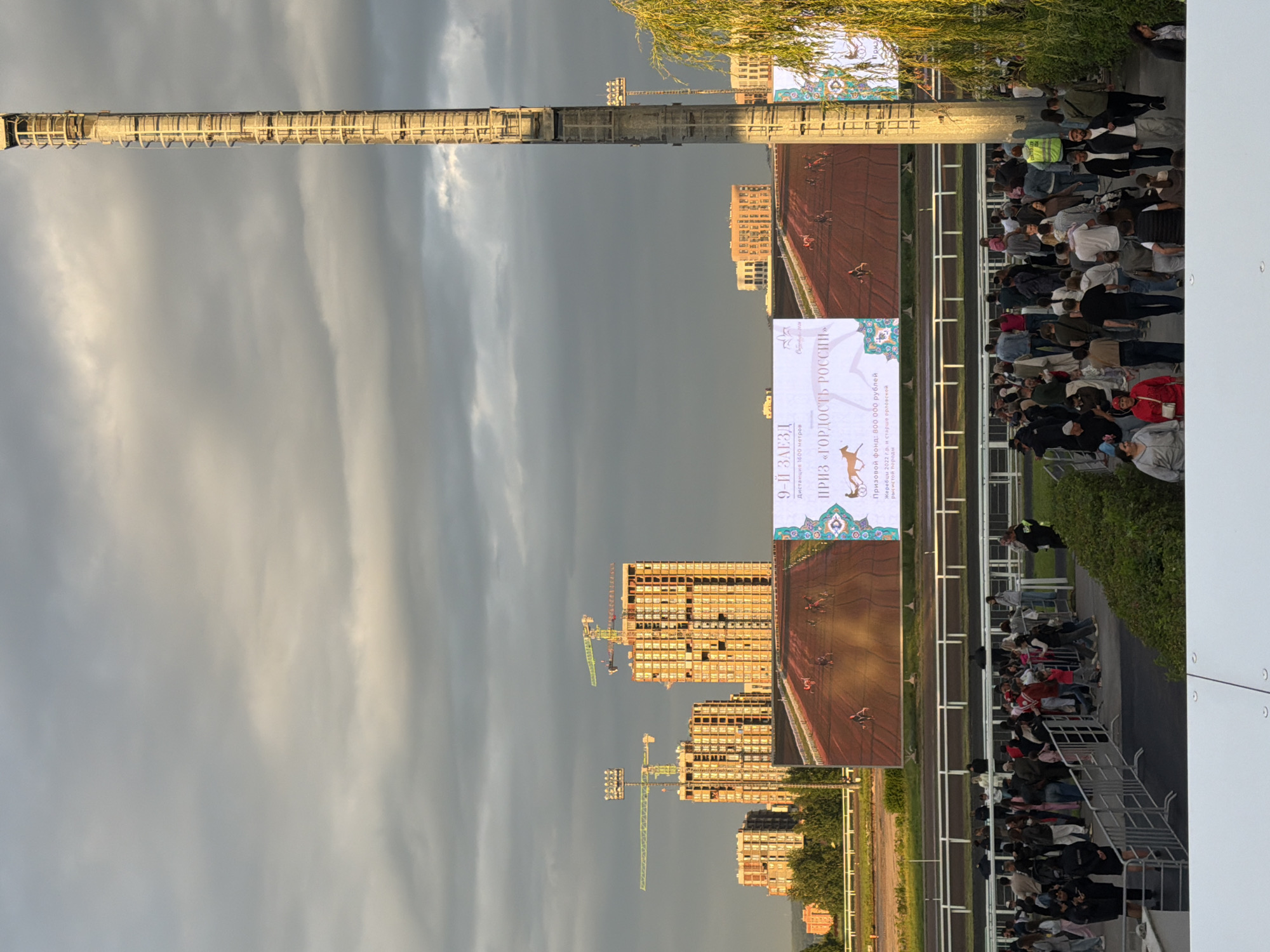 The image captures a wide-angle view of a horse racing track. In the foreground, a crowd of people, dressed in casual attire, are gathered behind a metal barrier, observing an event or waiting. The background features a large digital screen displaying a horse race in progress, with several horses and jockeys on a dirt track. Above the screen, construction of several tall buildings with cranes is visible, suggesting an ongoing urban development. To the right of the screen, a tall, industrial-looking structure, possibly a lighting tower, dominates the scene. The sky is overcast with dramatic clouds, and a weeping willow tree partially frames the right side of the image. The overall impression is a bustling outdoor event set against a backdrop of urban construction and a dynamic sky.