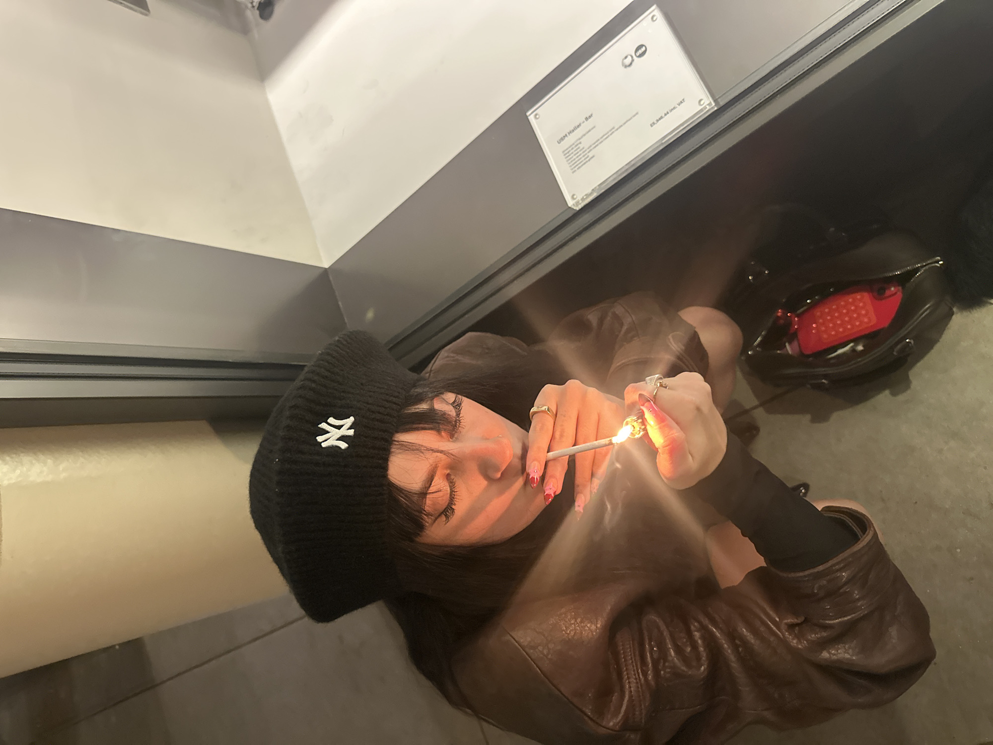 A female individual, viewed from an elevated perspective, is depicted lighting a cigarette. She has dark hair and wears a black knit beanie with a white New York Yankees logo, a brown leather jacket over a black long-sleeved top, and multiple rings. Her fingernails are painted pink with red tips. She holds a silver-filtered cigarette to her lips with her right hand while igniting it with a gold-colored lighter held in her left hand; the flame illuminates her face with an orange glow. A small nose piercing is visible on her left nostril. To her right, a black bag containing a red object is present on a dark floor. Adjacent to the individual, a display case or window features a white rectangular label that reads "USM Haller - Bar" and displays a price of "£5,968.44 inc. VAT". This label, combined with the pricing in British Pounds and the brand name "USM Haller," indicates the scene is located within a high-end furniture showroom or retail establishment for modular furniture, specifically in the city of London, United Kingdom.