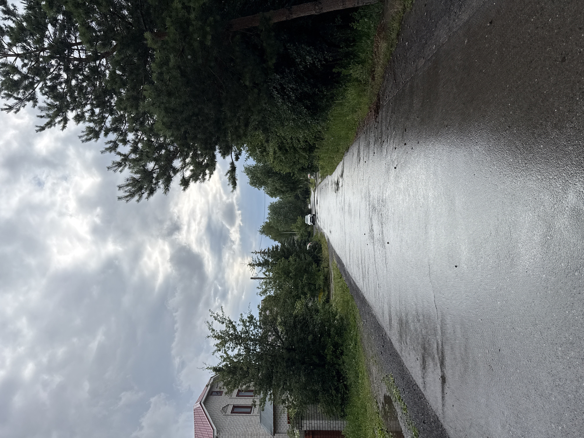 The image depicts a wet asphalt road running through a semi-rural area, likely in Kazan, Russia, under an overcast sky. Dense green trees line the left side of the road, with a two-story house visible in the foreground. The house has light-colored brickwork and a red-tiled roof. The road surface is reflective, indicating recent rain, and scattered dark spots suggest fallen leaves or debris. A few cars are visible in the distance on the road. The sky is filled with heavy, grey clouds, creating a muted and somewhat somber atmosphere. The overall impression is of a quiet, potentially post-rainy day in a suburban or village setting. No people are clearly visible, and no text is discernible in the image. The perspective is from a low angle, looking down the road and towards the sky.