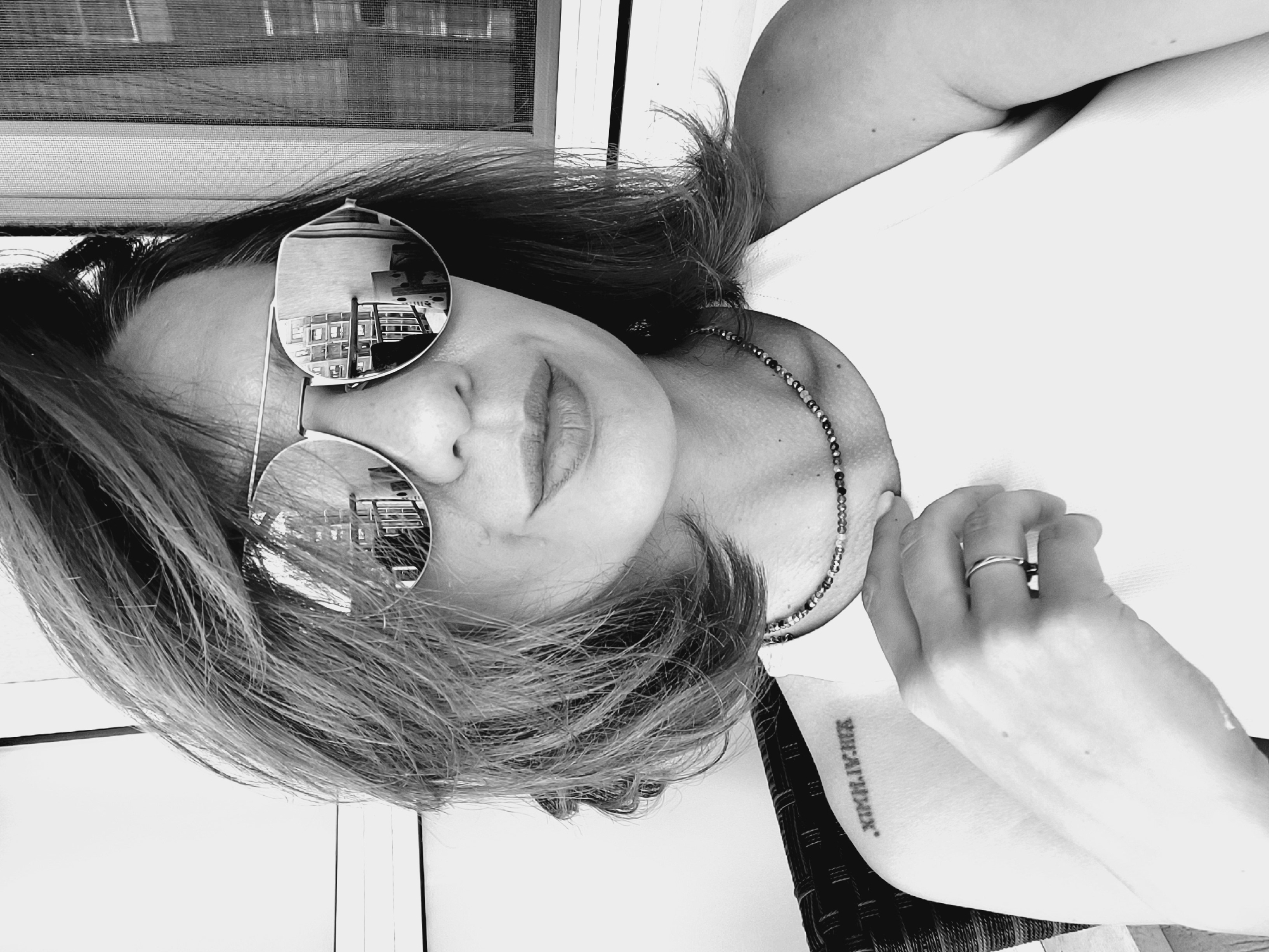 A black and white, close-up selfie shows a woman with short, dark hair and sunglasses. She is wearing a white tank top and a beaded necklace. A ring is visible on her left ring finger. A tattoo of Roman numerals is visible on her left shoulder. The background appears to be an outdoor setting, possibly a balcony or patio, with furniture and a window with a screen. The reflection in her sunglasses shows an indoor scene with shelves of books or objects. The location is not specified in the image.