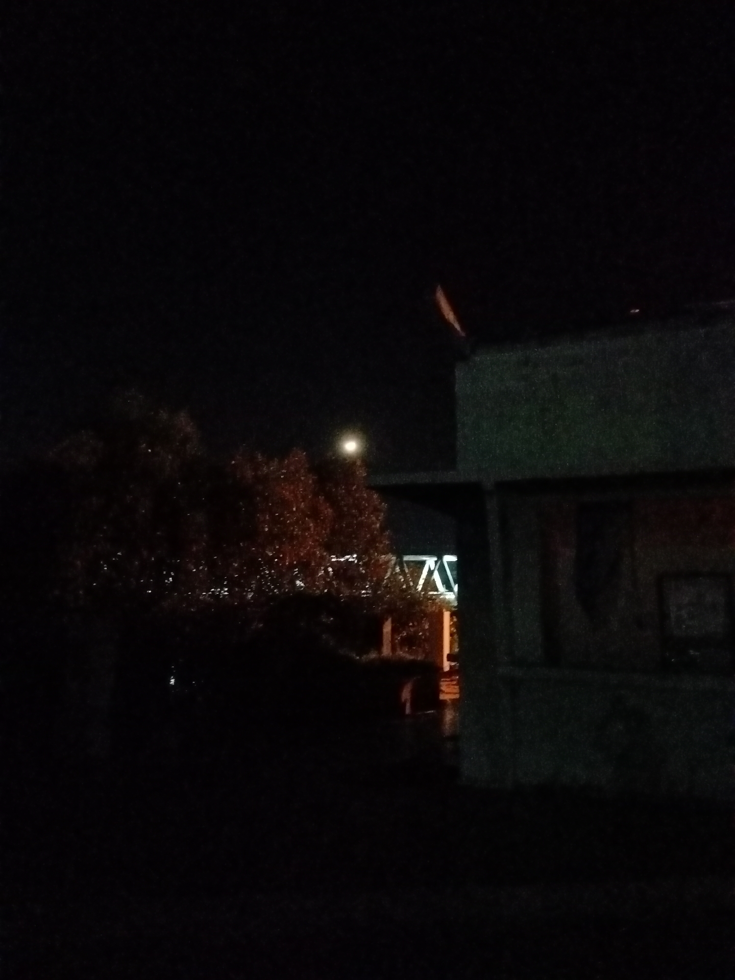 The image, taken at night in Lhokseumawe, Indonesia, shows a dark urban scene. In the foreground is a concrete structure, possibly a building or shelter. Behind it, a tree with illuminated leaves obscures a bridge structure with angular supports. A dim light source, possibly the moon or a streetlamp, is visible in the sky behind the tree. The foreground is shrouded in darkness, making it difficult to discern details. The scene suggests an urban environment with infrastructure and vegetation, captured under low-light conditions. No people are visible.