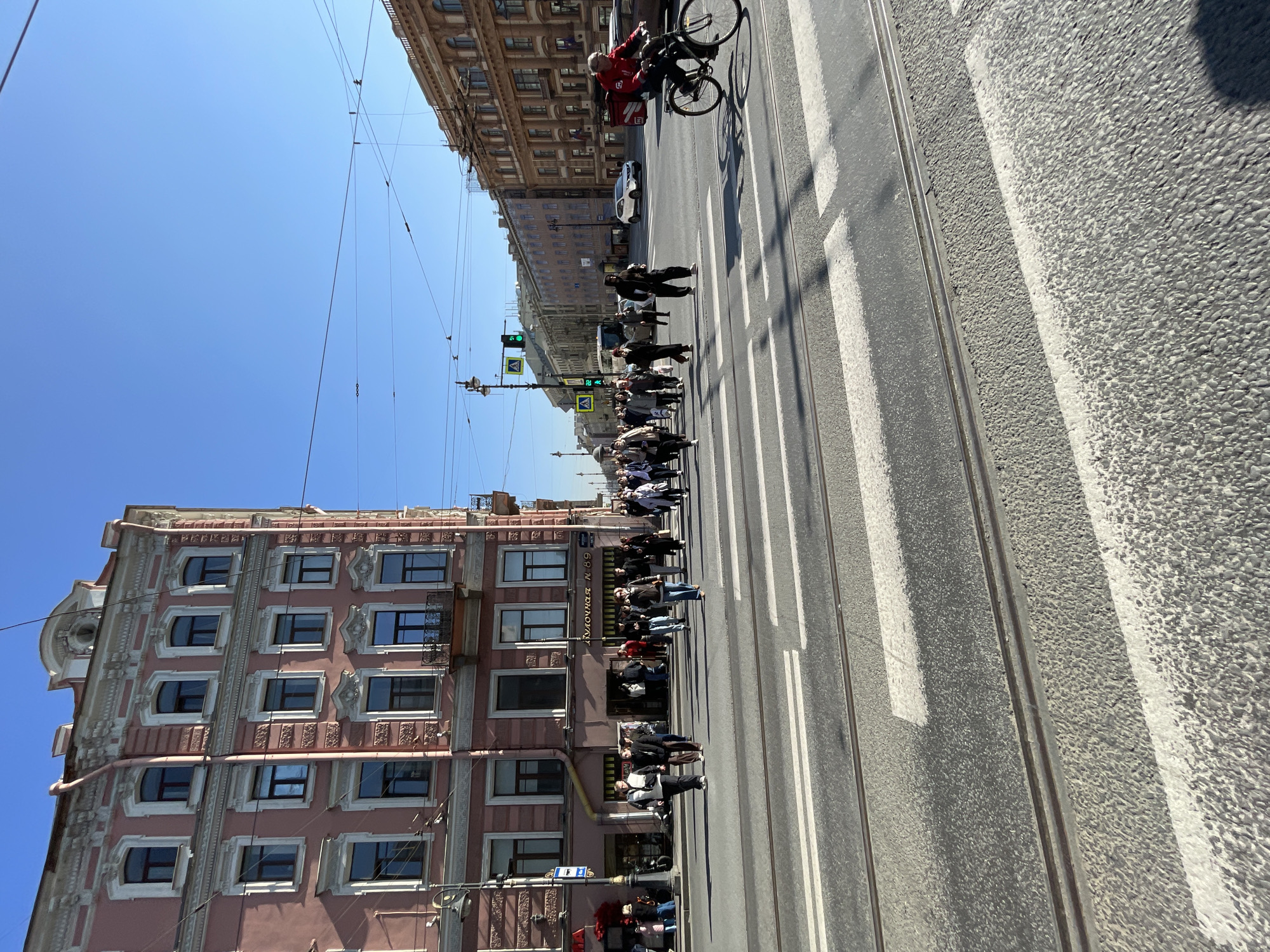 The image depicts a street scene in Saint Petersburg, Russia, viewed from a slightly elevated position. A pink building with multiple windows and architectural detailing is on the left, and a more modern building is visible across the street. A group of people is crossing the street on a crosswalk, while a cyclist is riding on the road. A tram line is visible along the street, parallel to the pedestrian crossing. Traffic lights show a green signal. Overhead electrical lines are seen against a clear blue sky. The scene appears to be taken during the daytime.