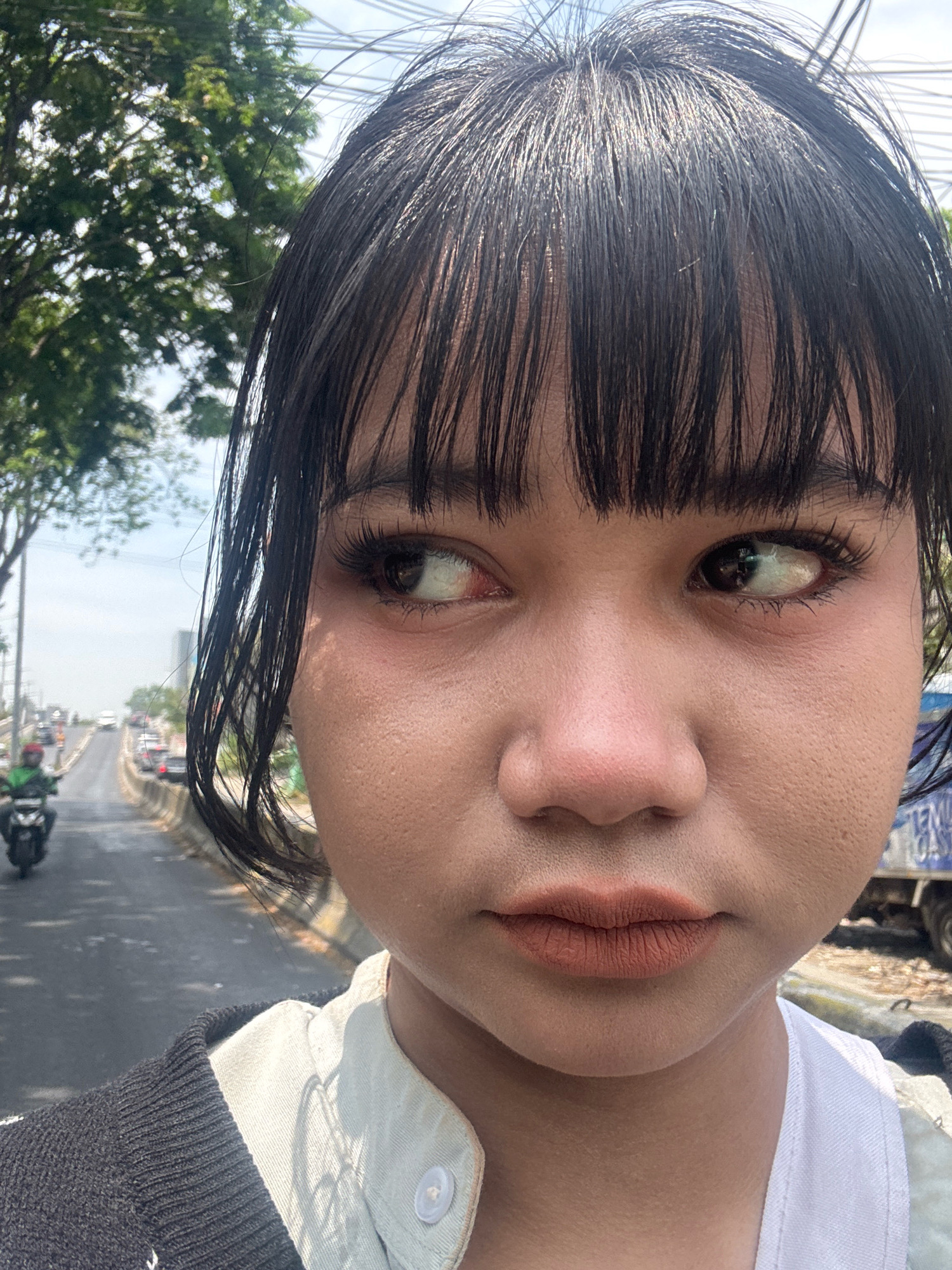 The image is a close-up selfie of a young woman with dark hair and bangs. She is wearing a white collared shirt and a dark sweater. Her makeup includes eyeliner and mascara on her eyes and matte lipstick on her lips. She is looking to her left. The background shows a street with traffic, including cars and a motorcycle. Trees and utility wires are also visible. The scene appears to be outdoors during the day. The location is identified as North Jakarta, Indonesia, implying an urban environment.