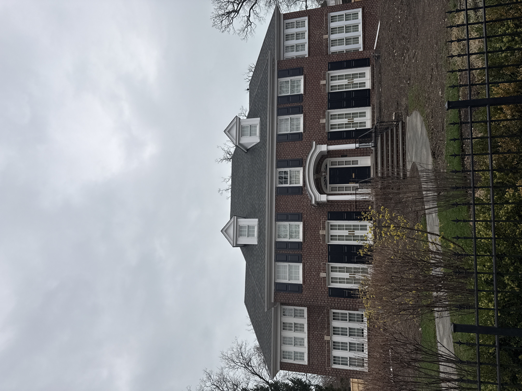 A three-story brick building stands against a cloudy sky. The building has a grey roof with three white dormers. Black shutters flank the white-framed windows. A black door is located centrally on the building's first floor, beneath a white arched portico. Steps lead up to the entrance. A black wrought iron fence borders the property. Bare trees and shrubs are visible in the foreground. The location is Winnetka, United States of America.