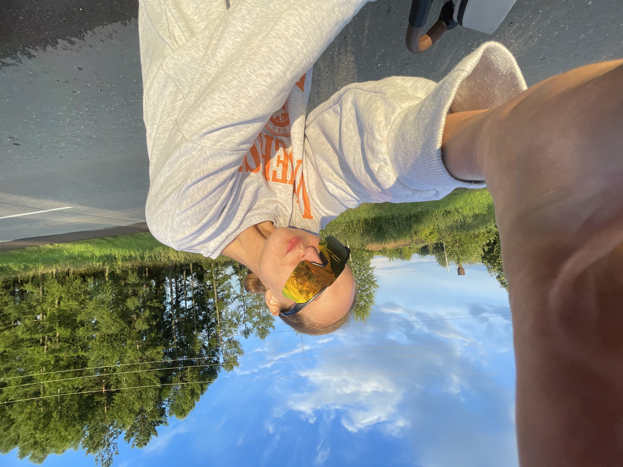 In the city of Podshivalovo, Russia, a person wearing large, reflective sunglasses and a light gray sweatshirt with orange lettering takes a selfie outdoors. The individual's arm extends towards the camera, partially obscuring the view. Behind them, a paved road runs horizontally, bordered by a grassy verge and a line of tall, green trees under a bright blue sky with scattered clouds. A single utility pole with wires is visible in the distance.