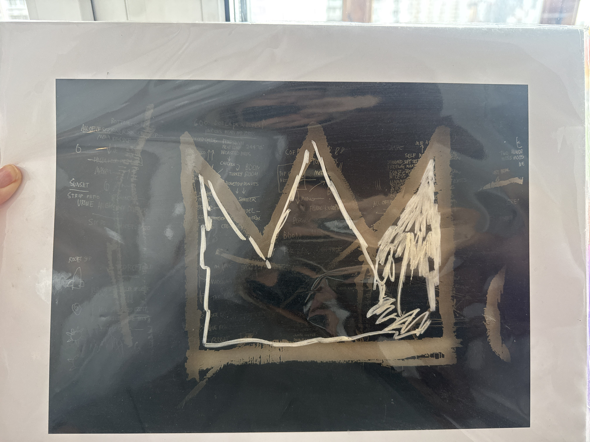 The image displays a framed piece of art, likely a print, covered in a protective plastic sleeve. The artwork itself features a prominent, stylized crown drawn in thick, light-colored paint against a dark, textured background. The crown has a rough, almost jagged appearance, with a few loose brushstrokes giving it a dynamic feel. Surrounding the crown are numerous scribbled lines, symbols, and text in the same light color.
The text appears to be a mix of words and numbers, some legible and some partially obscured or stylized. Phrases like "BOTTOMLESS," "ALL OF OF SERYONG RSP," "SUNSET," and "URINE HIGHWAY" are visible. There are also lists of items and phrases like "600 DOLLARS IN DINES," "VARIOUS MEARS AND PORTS," "PICCED CHEESE FRIED SALAMI," "CHICKEN BOOM," and "TURKEY BOOM." Small, simple drawings of objects, such as what might be a rocket ship, also dot the background.
The overall aesthetic is raw and expressive, characteristic of street art or neo-expressionism. The background's texture and the frenetic nature of the scribbled text suggest a chaotic or energetic subject matter. The plastic sleeve shows some reflections and smudges, indicating it is not a professional gallery setting, but rather a more personal display or perhaps a product awaiting sale. The lighting suggests it's an indoor scene with natural light coming from a window in the background, which is out of focus. There are no people directly depicted in the artwork. The context of Moscow, Russia is not visually represented in the image itself, but is provided as external information.