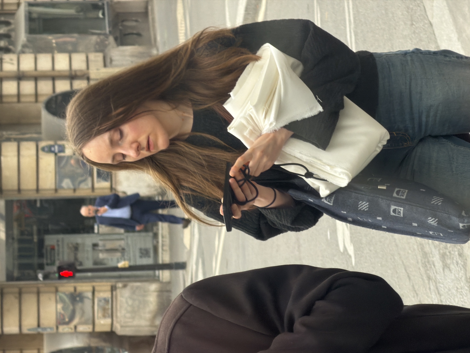 A daytime street scene in Paris, France, features a woman with long, light brown hair, wearing a black sweater and blue jeans, who is seated and holding a white folded textile, as well as a black cell phone and headphone wires in her hands. Beside her sits another individual, wearing a brown hoodie. The background includes a blurry view of a man in a suit walking past storefronts, traffic lights, and typical Parisian buildings.