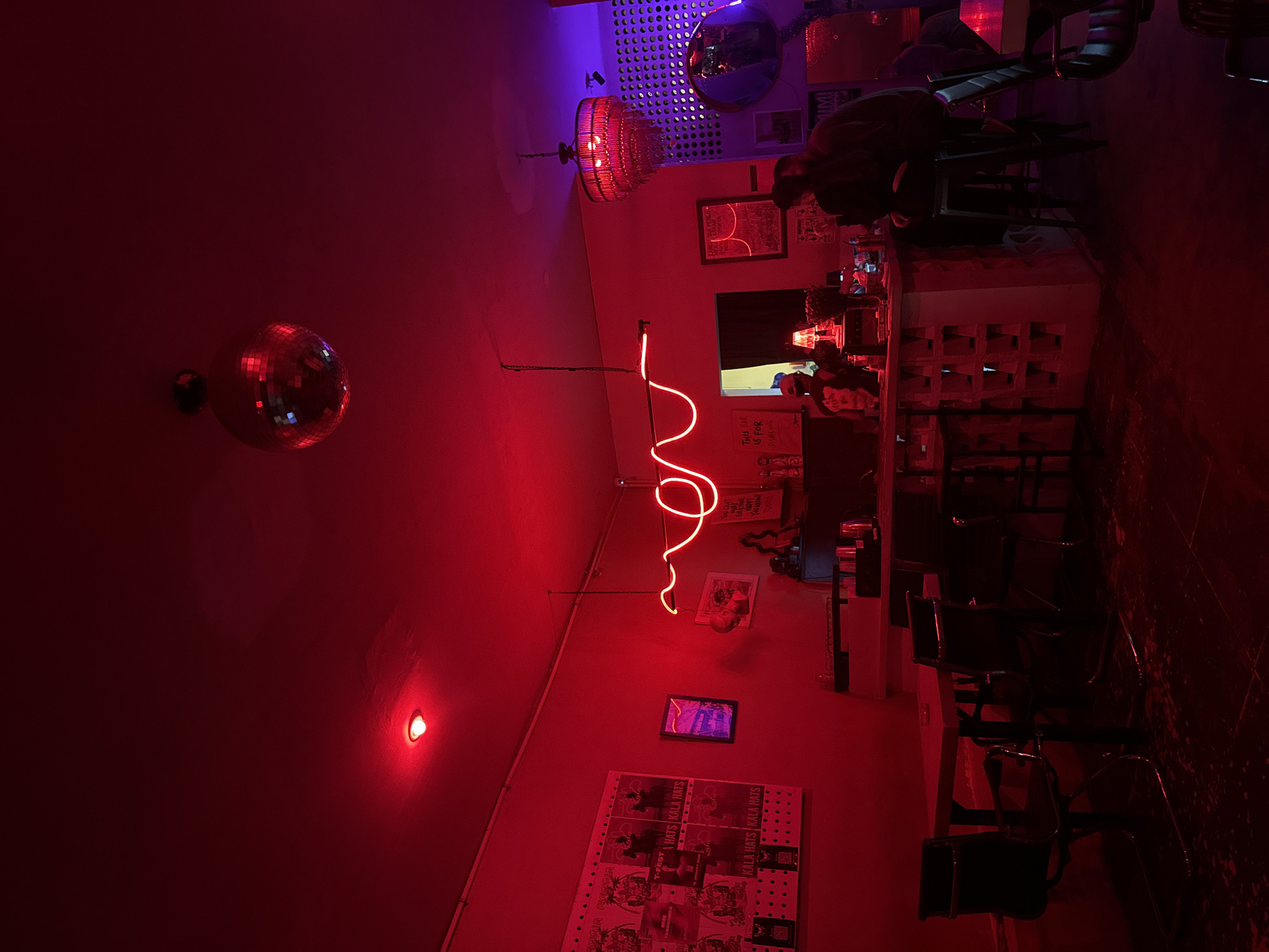 The image shows the interior of a bar in North Kuta, Indonesia. The scene is lit with red and purple lights, casting a reddish hue over everything. A disco ball hangs from the ceiling. A neon sign in the shape of a squiggly line is mounted on the wall, along with posters and framed pictures. People are seated at the bar, including one man sitting on a high chair. The bar itself is white, with shelving behind it. Bar stools are arranged along the bar. A chandelier hangs from the ceiling in the background.