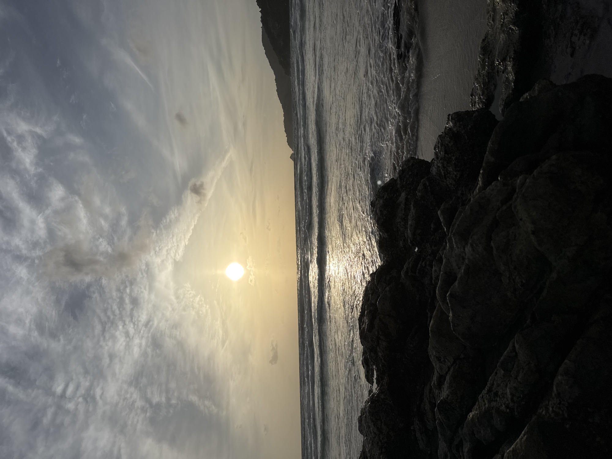 The image depicts a coastal scene at sunset, presumably near Patong, Thailand. In the foreground, dark, wet rocks are scattered across the shoreline. The ocean extends to the horizon, with moderate waves breaking and creating a foamy edge as they reach the shore. The sun, a bright orb, is positioned in the upper center of the frame, casting a warm, golden glow that reflects on the water's surface. Wispy clouds streak across the sky, illuminated by the setting sun. In the distance, a lush, green hillside slopes down towards the sea, suggesting a tropical environment. There are no people or discernible human activity visible in the image.