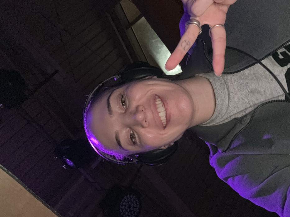 The image displays a smiling female individual in a close-up, upward-angled selfie taken within a dimly lit venue in Moscow, Russia. She wears dark over-ear headphones, a dark grey zippered jacket over a light grey high-necked shirt. Her left hand is raised towards the camera, forming a 'V' sign with her index and middle fingers. Multiple rings are visible on her left index and ring fingers, and a small tattoo is present on the side of her left index finger. The background features a dark, possibly wooden, ceiling structure with visible planks or beams. Several stage lighting fixtures are present; a multi-LED PAR light on the far left emits a distinct purple illumination that bathes the scene and the subject in a purple hue.