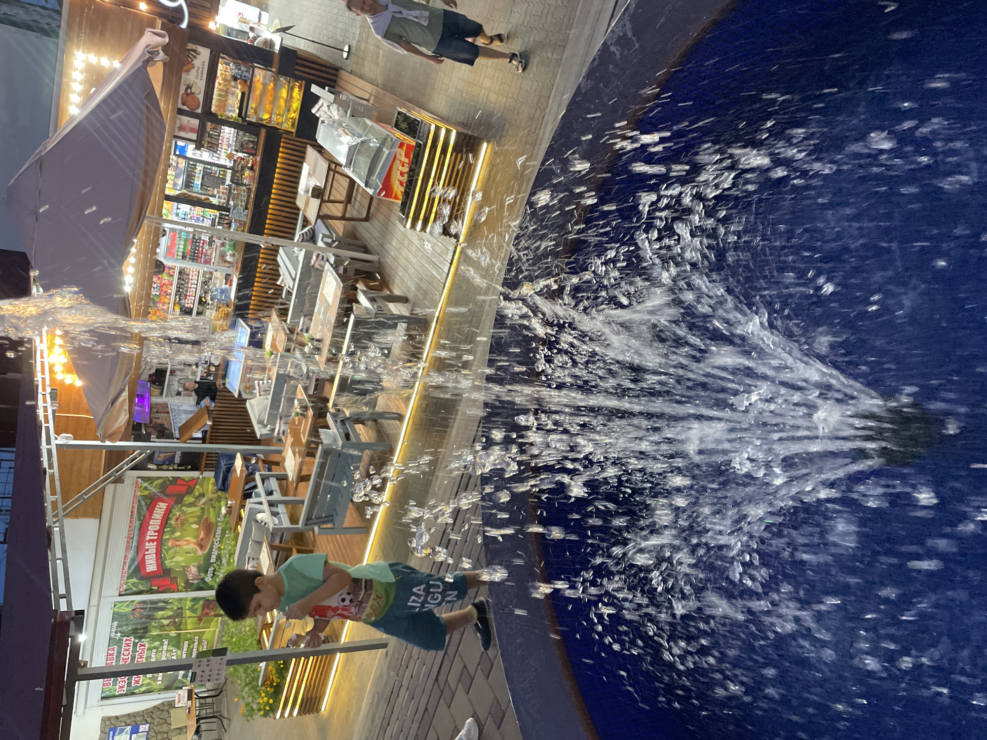 A fountain sprays water in the foreground, obscuring the view of an outdoor seating area with tables and chairs. A young boy in a green t-shirt and blue shorts stands to the left, holding a white object, possibly an ice cream cone. Behind him, a building with an awning and bright lights features signs with Russian text, one reading "ЖИВЫЕ ТРОПИКИ" (Live Tropics). A man in a light green shirt and dark shorts walks to the right of the frame. The scene appears to be a cafe or restaurant in a bustling area, likely in Khostinskiy, Russia, given the signage.