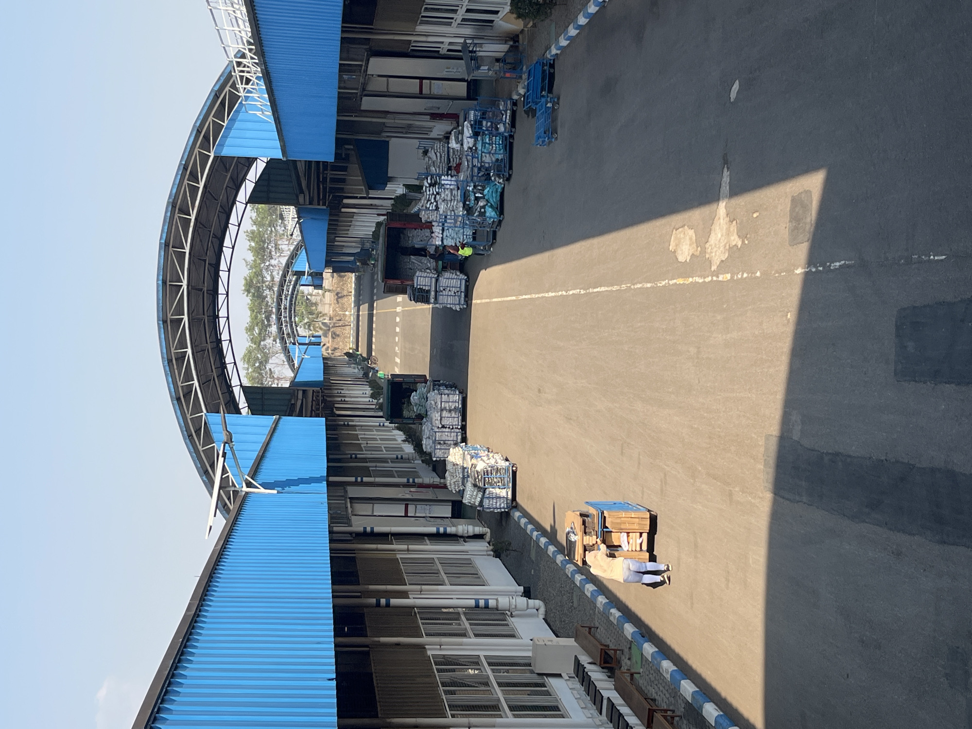 This is a high-angle, daytime photograph of an industrial facility, likely a warehouse or distribution center, in Pelang, Indonesia. The scene features a long, paved roadway running between two parallel buildings with bright blue corrugated metal roofs and sides. The buildings have white vertical pillars and numerous windows. Several open-top transport carts, some laden with goods wrapped in plastic, are positioned along the roadway.
In the foreground, a person wearing a light-colored shirt and blue jeans is pushing a cart filled with wooden boxes and other items. Further down the road, on the right side, a small teal-colored vehicle, possibly a forklift or a pallet jack, is visible near stacks of goods on carts. On the left side, further into the distance, a red truck is being loaded or unloaded, with people in the vicinity and more carts with goods. A person in a yellow vest is also visible near the truck.
The roadway is marked with a white line down the center, and the edges are bordered by a blue and white striped curb. Benches are lined up along the left building's exterior. Trees are visible in the background, beyond the structures. The sky is clear and light blue, indicating a sunny day. The overall impression is one of industrial activity and logistics.