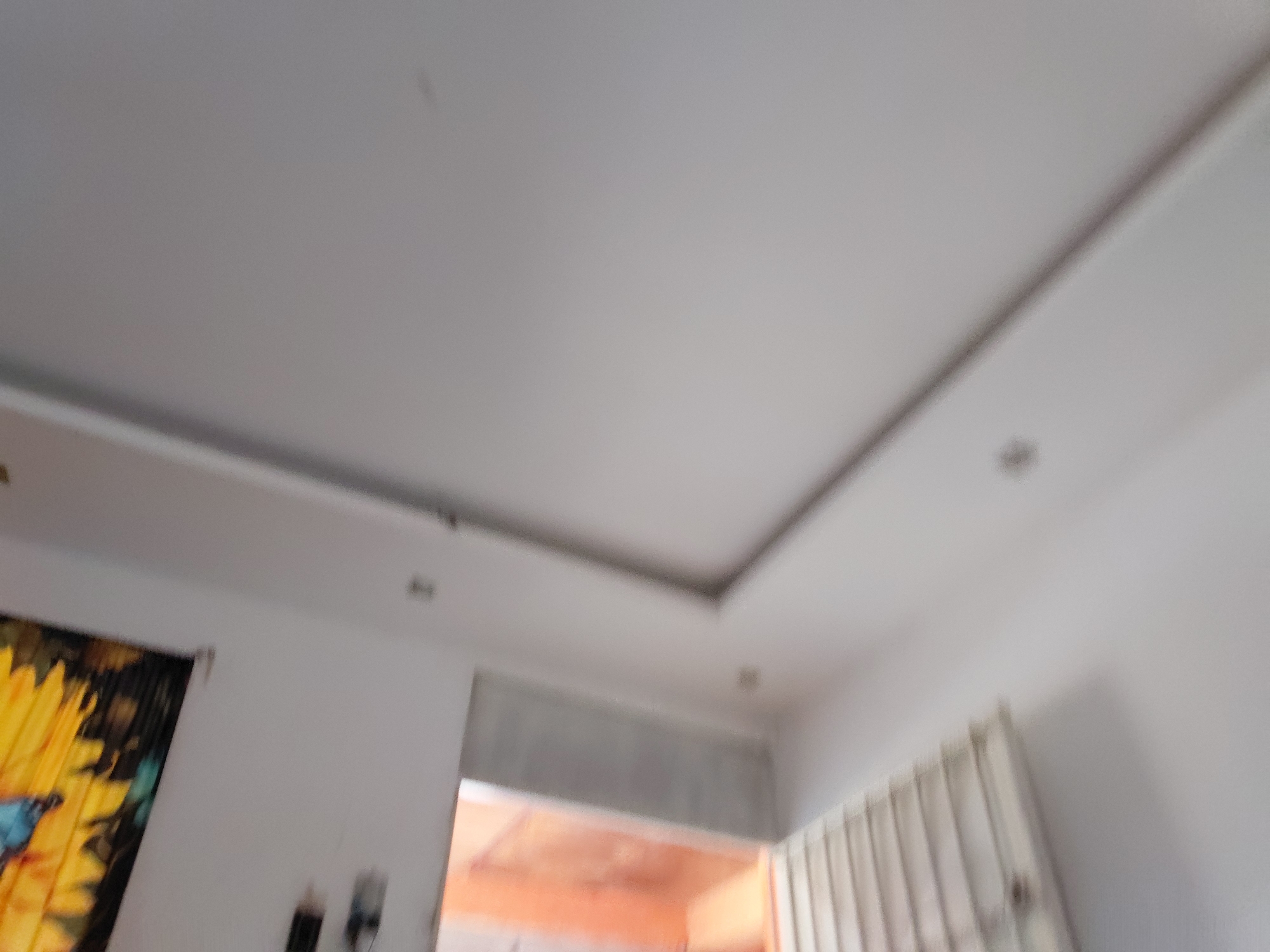 The blurry, upward-angled image captures the interior of a room in Santa Ana, Venezuela, focusing primarily on a white false ceiling. The ceiling features a multi-tiered design with a recessed rectangular section, likely intended for accent lighting, though no lights are visibly illuminated. Several small, dark specks, possibly dust on the camera lens or minor marks on the ceiling, are visible across the white surface.
On the left side of the frame, a partial view of a vibrant tapestry or curtain with a large yellow sunflower and blue design is visible. Below it, indistinct dark objects are partially seen against a white wall. Towards the right, a section of vertical blinds or a similar slatted structure is present. In the background, a brightly illuminated doorway or archway leads into another area, casting warm, reddish-orange light into the room.
No people are present in the image, and no specific activity is taking place. The lighting suggests it is daytime. There is no discernible text.