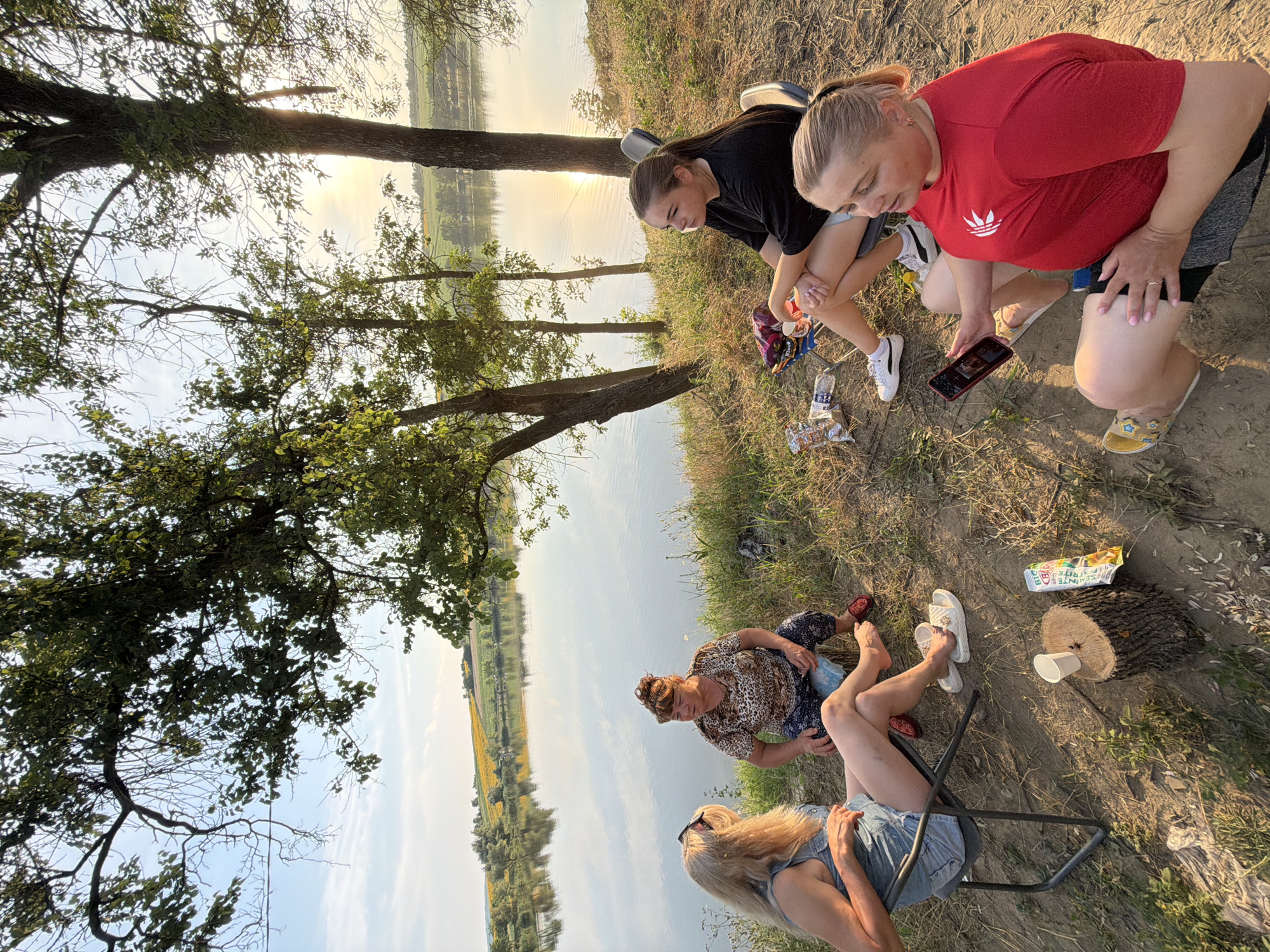 In Pănășești, Moldova, four women are gathered near a calm body of water, possibly a lake or river, under the shade of trees. The sun is setting, casting a warm glow on the scene. One woman, with blonde hair, sits in a camping chair, wearing a denim outfit and sunglasses. Next to her, another woman, with brown hair tied up, sits on a tree stump, dressed in a patterned top and dark pants. A young woman with dark hair in a ponytail is seated in a camping chair, wearing a black t-shirt and shorts. The fourth woman, with blonde hair pulled back, is in the foreground, wearing a red t-shirt and shorts, holding a mobile phone. Scattered around them are snacks, drinks, and personal belongings. The landscape features rolling hills in the background with a field of sunflowers visible, reflecting the golden hour light.