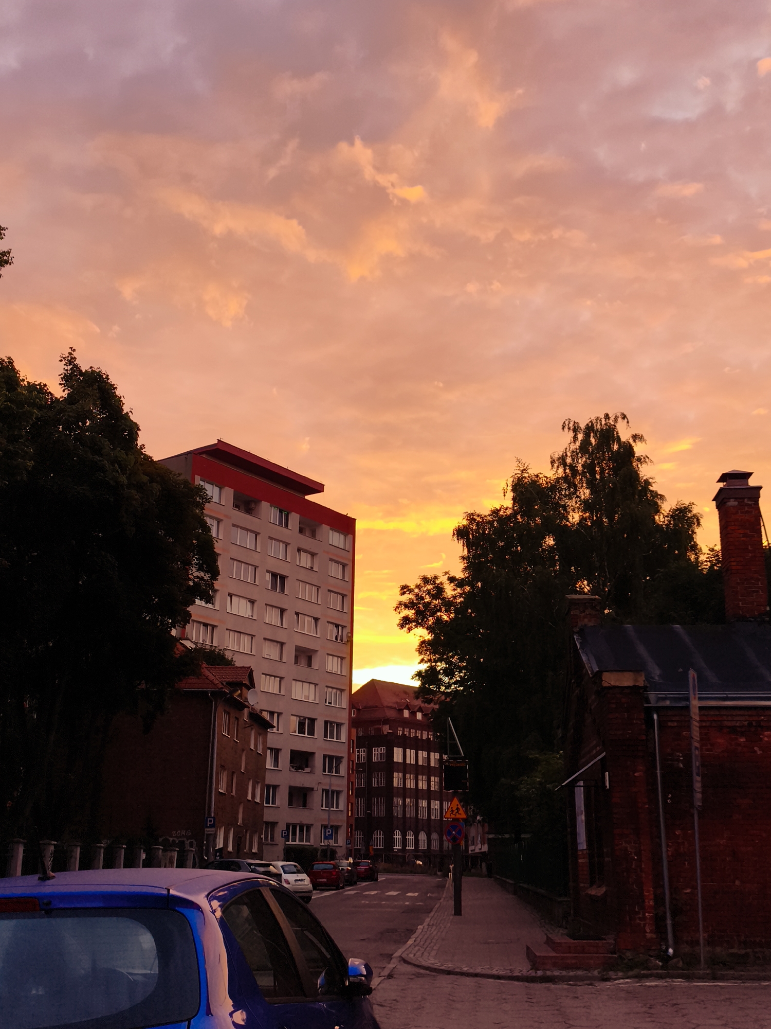 The image depicts a street scene at sunset in the Wrzeszcz district of Gdańsk, Poland. The sky is filled with vibrant orange and pink clouds, characteristic of a sunset. In the foreground, the hood and roof of a blue car are visible on the left. The street is paved with cobblestones.
Midground features a multi-story apartment building with light-colored walls and numerous windows, topped with a red roofline. To its left, a dark green tree partially obscures the building. Across the street, another, older brick building with a dark roof and a red brick chimney is visible on the right. This building is partially silhouetted against the bright sky.
Further down the street, more apartment buildings with many windows are visible, stretching into the distance. Traffic signs and streetlights are present along the sidewalk. The overall impression is of an urban environment during a picturesque sunset.