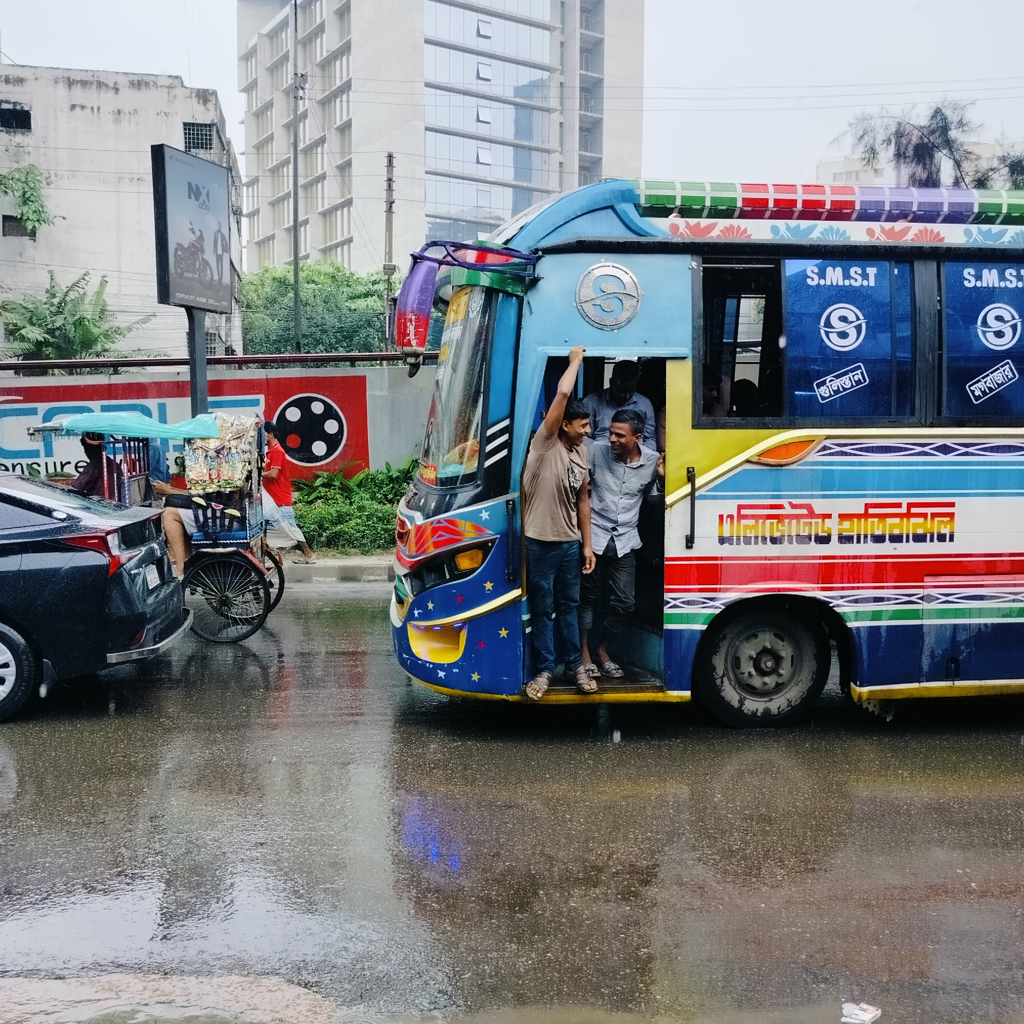 This is a scene from Dhaka, Bangladesh, on a rainy day. The image is dominated by a brightly colored, decorated bus with its door open, revealing passengers. A man is standing in the doorway, holding onto the top frame and smiling. Another man, dressed in a light-colored patterned shirt, is standing just inside the bus entrance, also smiling and looking towards the first man. Several other passengers are visible inside the bus. The bus itself is adorned with a colorful stripe design and stars.
To the left of the bus, a black car is parked, partially in the frame. Next to the car is a three-wheeled rickshaw, also covered by a light blue canopy. The rickshaw appears to be loaded with goods. A person in a red shirt is walking away from the rickshaw.
The ground is wet and reflects the surrounding environment, indicating recent rain. In the background, there are buildings, one of which is a modern high-rise with a grid-like facade. A billboard for "HONDA NX 200" is visible on the left side of the image, mounted on a pole. The overall impression is a busy urban street scene during a downpour.