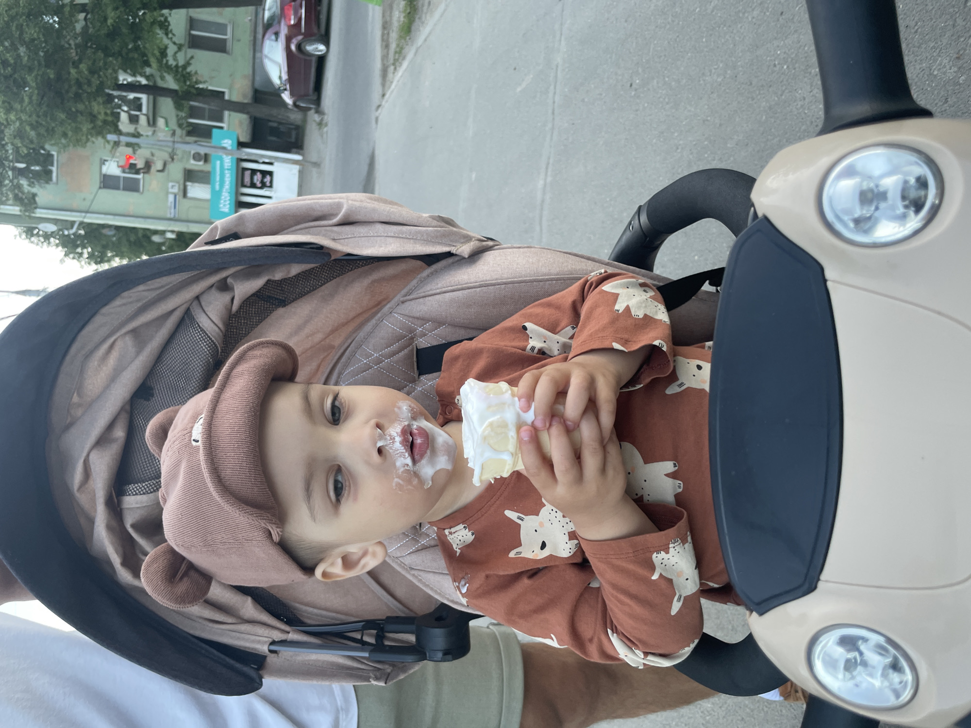A young child sits in a stroller, holding an ice cream cone. The child is wearing a brown shirt with animal designs and a brown hat with small bear ears. The child's face is covered in ice cream. The stroller is beige and has a dark gray hood. The background shows a city street with buildings and a car. A sign is visible on one of the buildings. Based on the building signage and architecture, the location can be identified as Izhevsk, Russia.