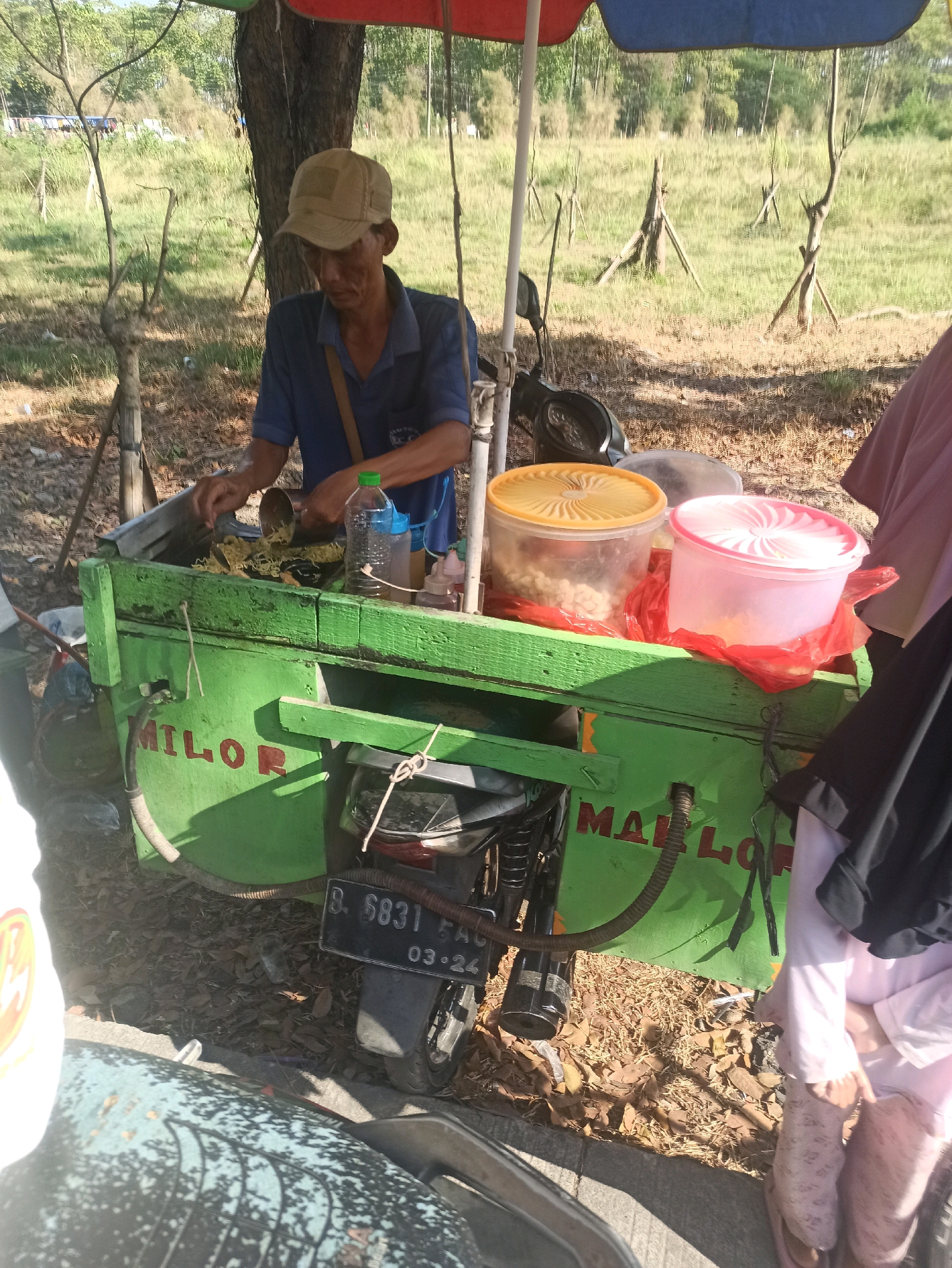 A man wearing a tan baseball cap and a blue shirt is standing behind a bright green food cart. The cart is mounted on a motorcycle, and a portion of the motorcycle's rear fender and license plate are visible, showing the text "B. 6831 FAQ 03.24". The man is holding a metal scoop and appears to be serving food from a large metal container on the cart.
In front of him are two plastic containers with lids, one yellow and one pink, containing food items. To his right, a person wearing a pink hijab and pink pants is visible, partially obscuring the cart. The cart has the red lettering "MAILOR" painted on its side. The background shows a grassy field with scattered trees and branches. The scene suggests a street-side food vendor operating in a somewhat rural or undeveloped area, likely Telagamurni, Indonesia, given the license plate format. The ground is covered with dry leaves and dirt.
