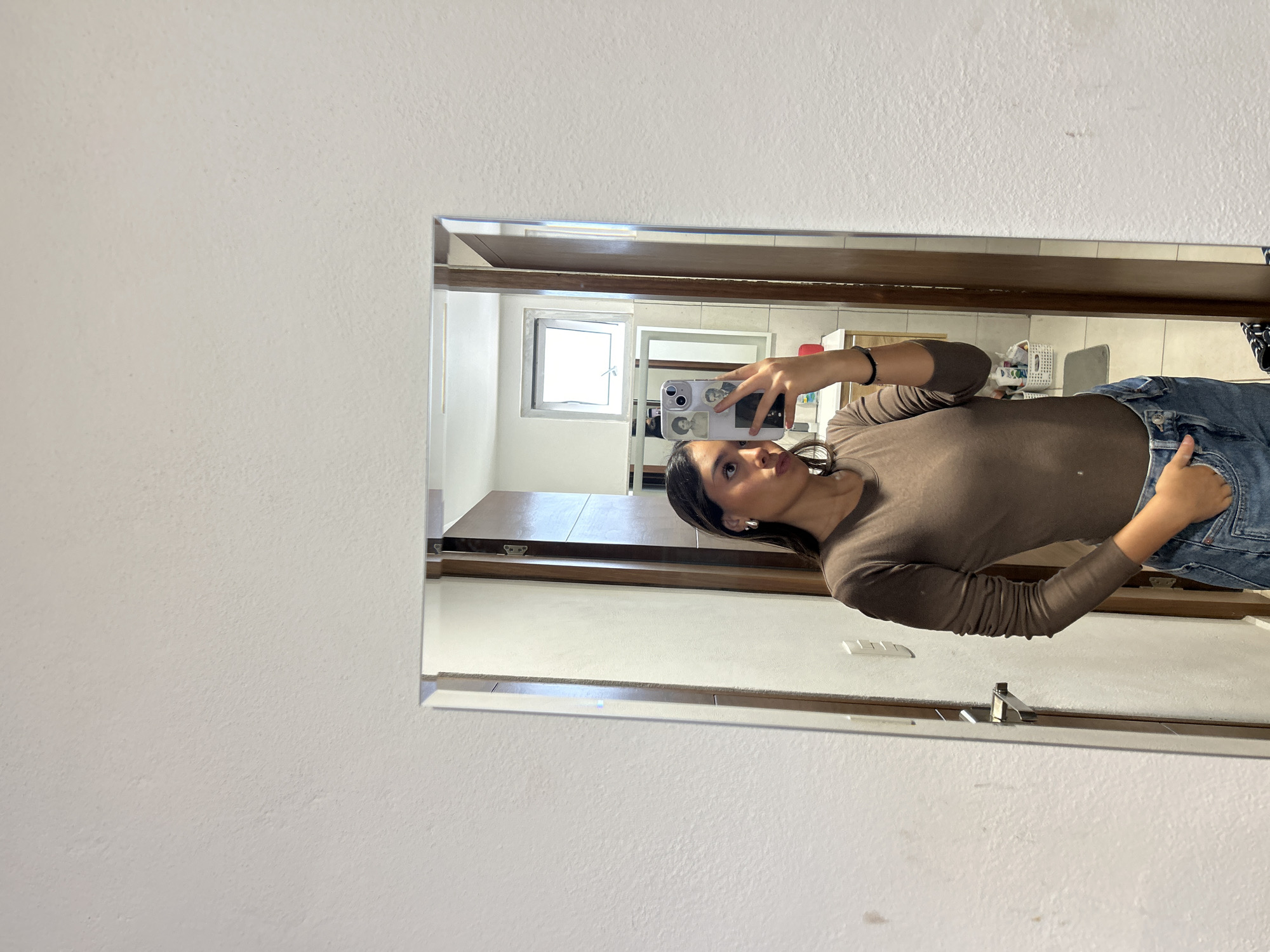 A woman takes a selfie in a mirror. The location is Unknown, Unknown. The woman has dark hair and is wearing a brown long-sleeved shirt and jeans. She is holding a smartphone in her hand. The mirror reflects a bathroom with a window, sink, and other objects.