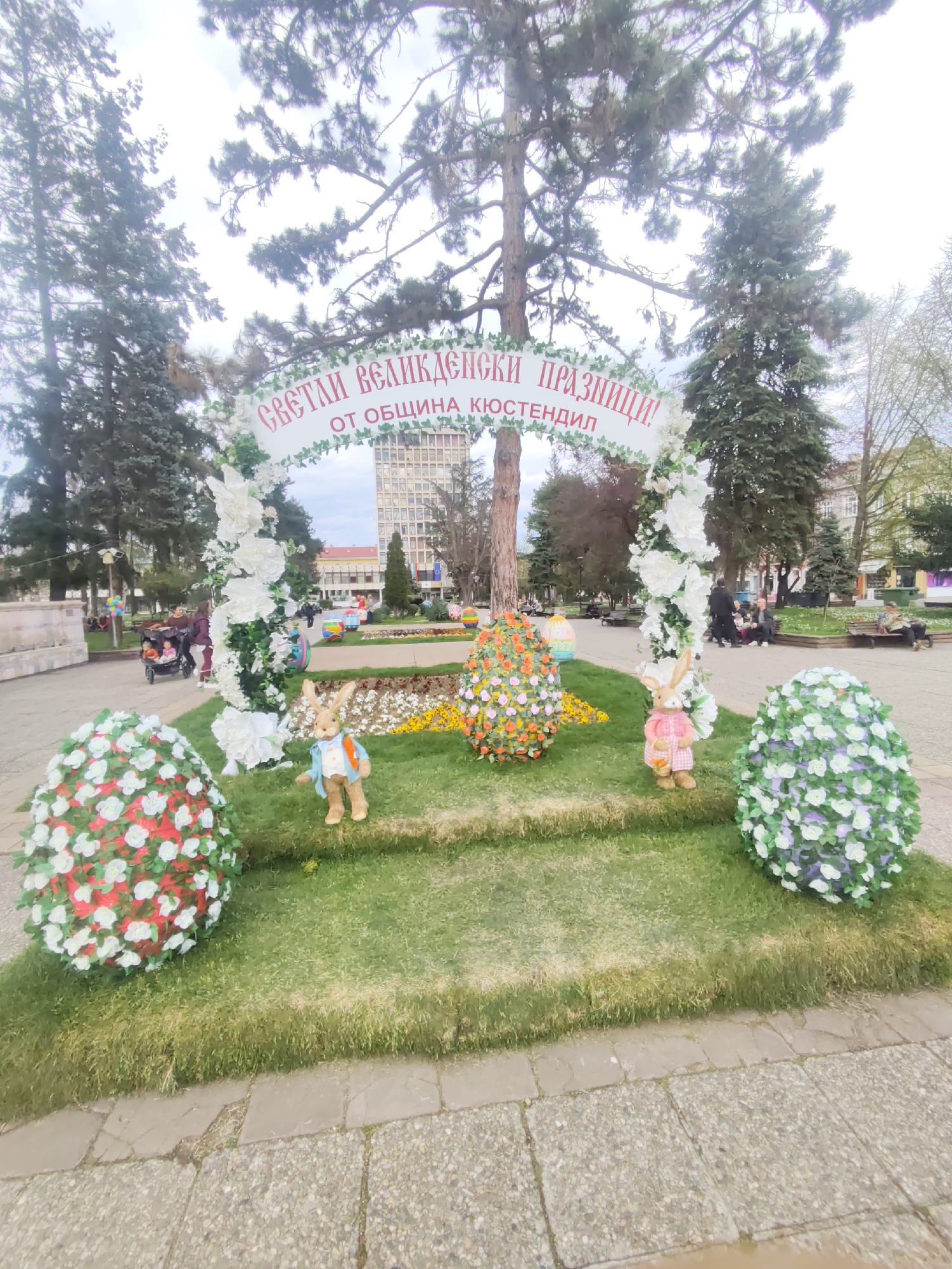 The image shows a spring festival setup in the city of Kyustendil, Bulgaria. The scene is located in a public square or park. The centerpiece is a decorative display celebrating Easter. It consists of a white arch covered in white flowers with a banner that reads "СВЕТЛИ ВЕЛИКДЕНСКИ ПРАЗНИЦИ! ОТ ОБЩИНА КЮСТЕНДИЛ!" (Bright Easter Holidays! From the Municipality of Kyustendil!).
Beneath the arch are three large Easter egg decorations made of flowers, each a different color scheme. Two stuffed bunny figurines are placed between the egg decorations. The setup is on a raised platform covered in grass.
In the background, other people are in the park, including individuals sitting on benches and a person pushing a stroller. Trees surround the area, and a tall building can be seen in the distance. The weather appears overcast. The ground in the foreground is paved with stone tiles.