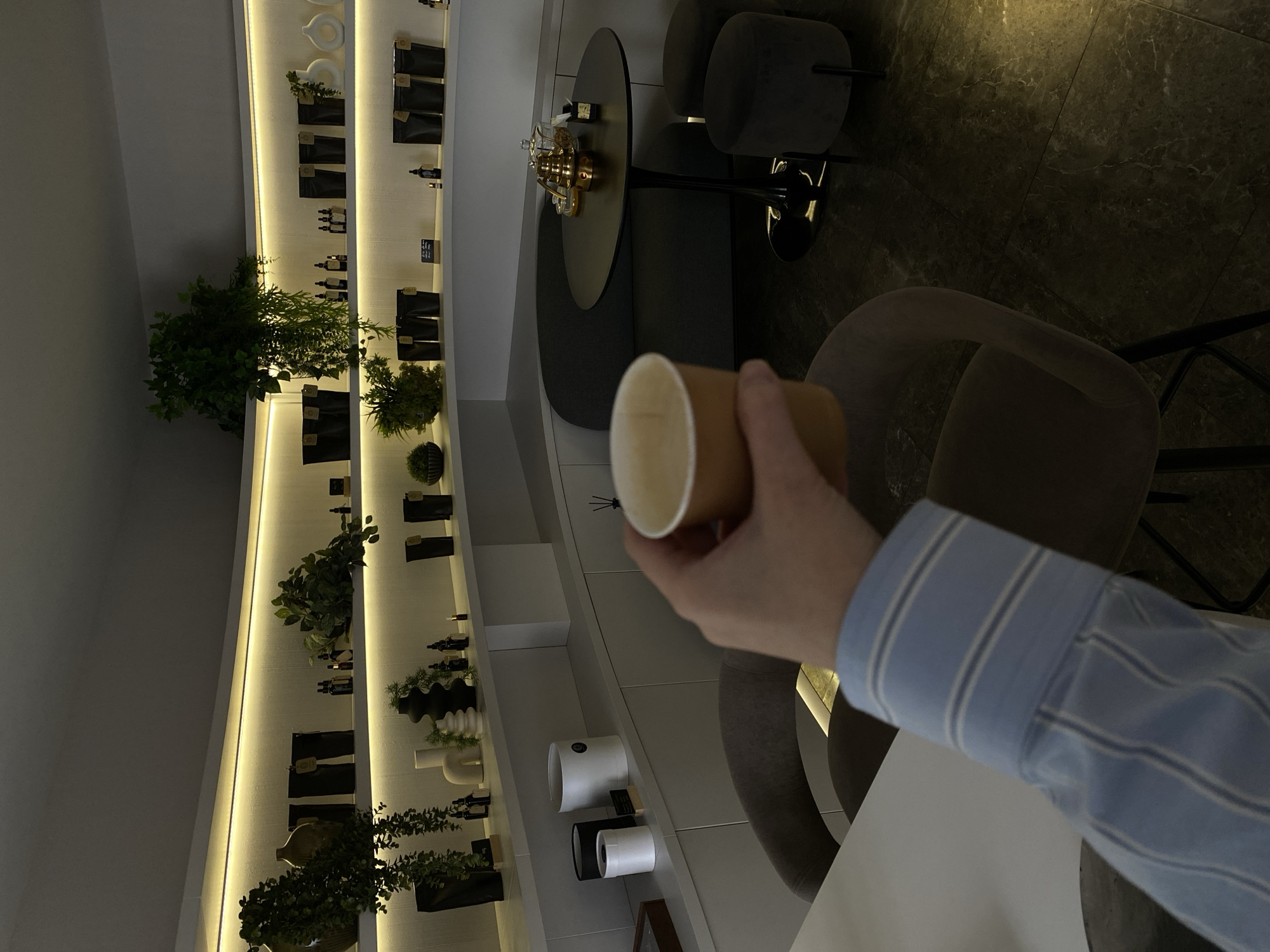 The media file captures an indoor scene in Votkinsk, Russia, depicting a modern establishment such as a cafe, lounge, or a retail space specializing in artisanal products. The image is taken from a first-person perspective, showcasing a person's right hand holding a brown disposable paper cup with a white interior. The hand wears the cuff of a light blue striped shirt.
The background is dominated by a long, multi-tiered shelf display along a wall, illuminated by warm LED strip lighting. The shelves feature various items including small potted plants or artificial greenery, numerous small bottles (possibly cosmetics or essential oils), and black pouches or bags. Decorative elements such as white ceramic rings, a black wavy vase, a beige vase, and two cylindrical containers (one white, one black) are also arranged on the shelves.
Towards the right side of the image, a seating area is visible with a round black table, adorned with a golden, intricate decorative object resembling a small ship. Dark grey upholstered chairs or stools, and a matching upholstered bench or booth seating, surround the table. The floor is composed of dark grey or black tiles with a subtle marble-like pattern.
The scene suggests a relaxed atmosphere, likely a place for customers to browse products or enjoy a drink. The artificial lighting creates an inviting ambiance. There is no clearly legible text visible on any of the objects or labels within the frame.