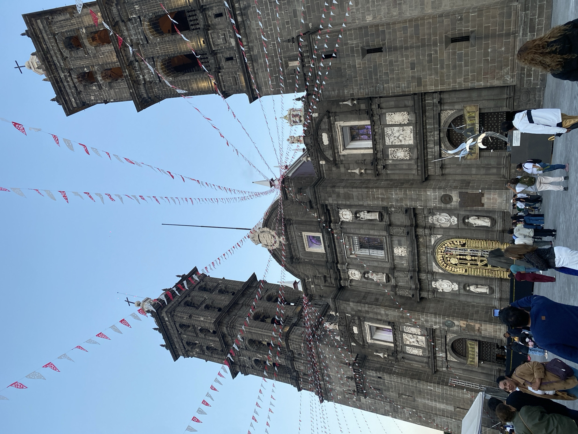 The image depicts the Puebla Cathedral in Puebla, Mexico. The cathedral is a large, gray stone structure with two tall bell towers adorned with crosses. Decorative paper banners with triangular shapes are strung between the towers and the central part of the building. The sky is a clear blue. Several people are present in the foreground, some appearing to be tourists or visitors observing the cathedral. An art installation, sculpture, or modern artwork is located in front of one of the cathedral's entrances.