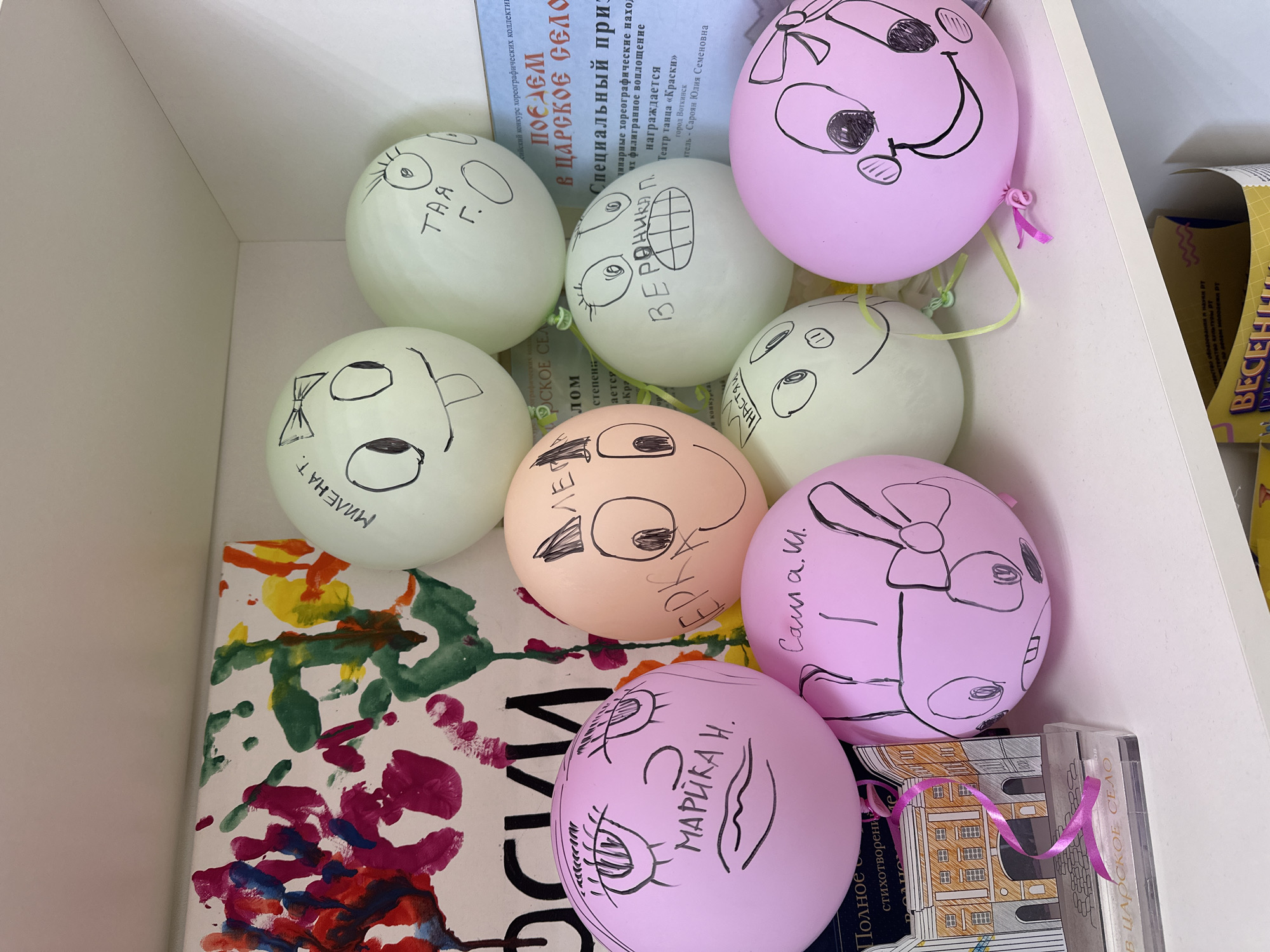 The image shows a collection of colorful balloons with hand-drawn faces and names on them, placed on top of books and other items inside a white cabinet or shelf. The balloons are light green, pink, and peach-colored. The names written on the balloons include "Taya", "Veronika", "Milena", "Erika", "Sasha", and "Marika". One of the books is partially visible and appears to be related to the city of Tsarskoe Selo. Another is a children's artwork painting of red, green, yellow and blue paint on white canvas. The image also shows a poster or flyer mentioning "Teatr tanca 'Kraski' gorod Votkinsk", indicating a dance theater located in the city of Votkinsk, Russia.