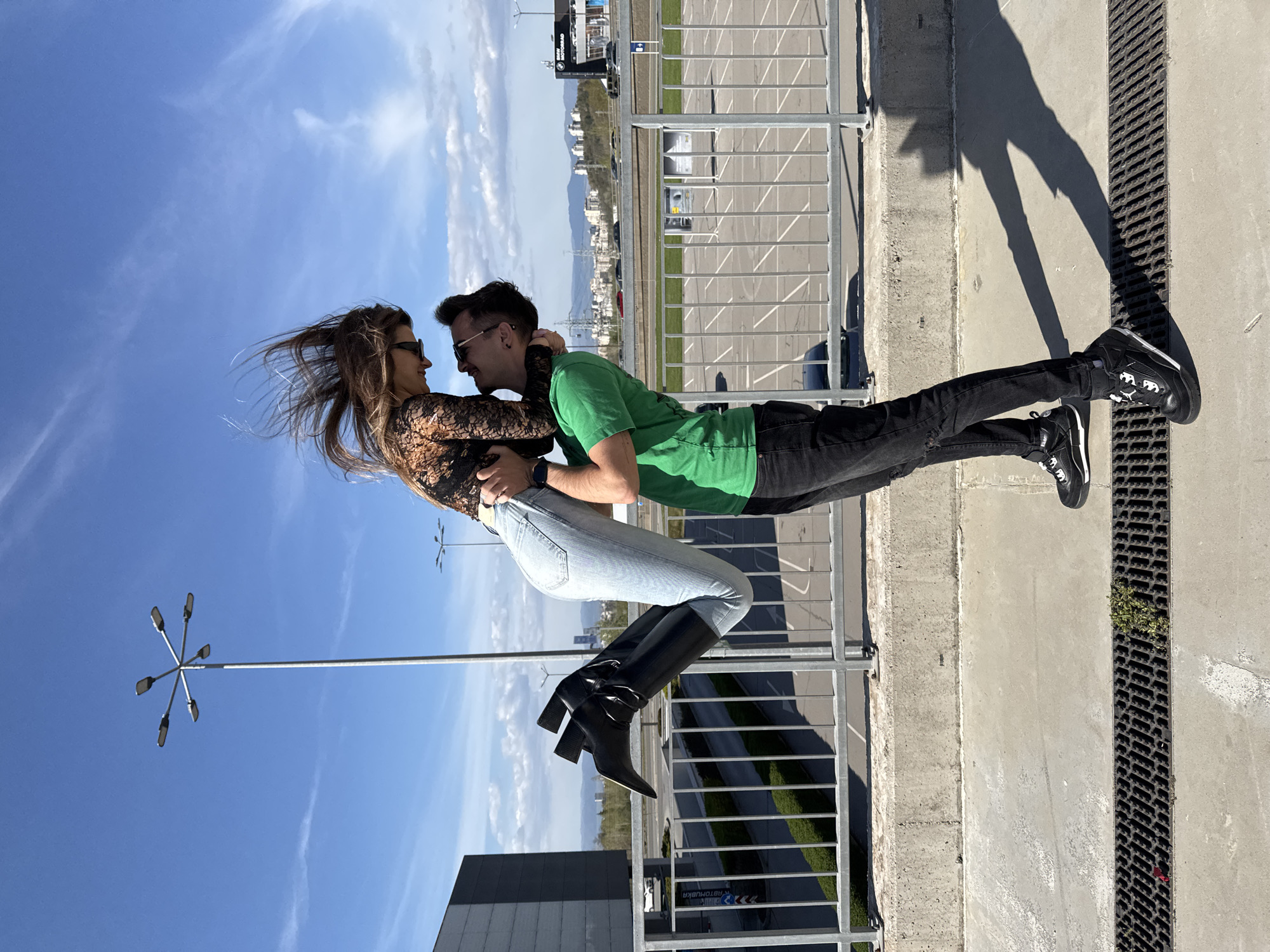 The image portrays a joyful and affectionate moment between a young couple in an urban outdoor setting, likely Sofia, Bulgaria, under a bright, sunny sky. The photo is taken from a slightly low angle, emphasizing the expanse of the sky.
In the center, a man and a woman face each other in a dynamic embrace. The man, on the right, wears a green short-sleeved t-shirt, dark jeans, and black sneakers with white details. He has short dark hair and sunglasses. The woman, on the left, is dressed in a black lace long-sleeved top, light blue skinny jeans, and tall, pointed-toe black high-heeled boots. Her long, dark hair flows dramatically around her head, suggesting movement or a strong breeze. Both individuals are looking at each other, conveying emotions of happiness and intimacy. The woman appears to be lifted or jumping slightly, adding to the playful nature of the pose.
The background features an expansive concrete area, possibly a parking lot or plaza, with a metal barrier or fence running horizontally across the mid-ground. Beyond the fence, urban buildings and a distant mountain range are visible under a clear blue sky with scattered wispy white clouds. A large billboard with the "BMW" logo is prominent above the man's shoulder. Modern streetlights with star-shaped light arrays are also visible against the sky.
The immediate foreground shows a concrete ground surface with a metal drainage grate running along the right side. Strong, distinct shadows of the couple are cast on the ground to their left, indicating direct sunlight. Partially visible on the ground below the woman's feet are signs with Cyrillic text, including "АВТОМИВКА" (Car Wash) and "АВТОПАРК" (Car Park). The scene is vibrant, capturing a moment of romance and energy during daytime.