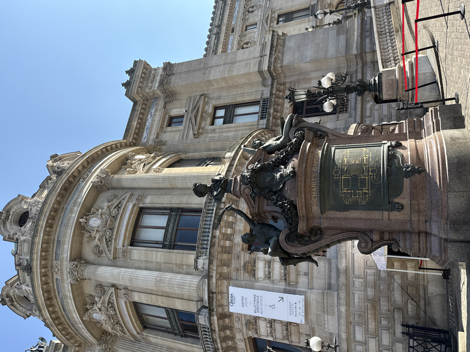 The image captures the exterior of the Opéra Garnier (Palais Garnier), located in the 9th arrondissement of Paris, France, under a clear blue sky. A large, ornate monument dedicated to Charles Garnier dominates the foreground. This monument features a polished granite or marble base inscribed "1825 CHARLES GARNIER" and includes a bronze plaque depicting an architectural plan. Above the base, bronze sculptures depict a male bust at the apex, flanked by allegorical figures including a nude male reaching upwards and a semi-reclined figure. The Opéra Garnier building, constructed of light-colored stone, rises behind the monument, showcasing its grand Neoclassical-Baroque architectural style. Key features include a prominent rounded corner with multiple ornate windows, Corinthian columns, pilasters, balustrades, and intricate decorative carvings such as garlands, putti, and medallions. A white banner hangs on the building's facade, displaying "OPERA," "VISITES," "BILLETERIE SPECTACLE," and "BOX OFFICE" text in French and English, along with directional arrows and the website "OPERADEPARIS.FR." Ornate black street lamps with multiple globes are positioned on the paved surface to the left and right of the monument. Red rope barriers on black stanchions guide pedestrian traffic in the lower right portion of the scene. No individuals are visible.
