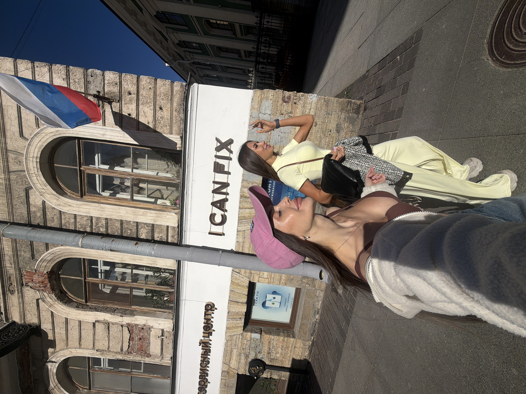 Two women are walking on a sidewalk in front of a building in Saint Petersburg, Russia. The building has arched windows and a weathered facade. A Russian flag is visible hanging from the building. One woman is wearing a pink baseball cap and a dark top, while the other is wearing a light yellow outfit. The sidewalk is made of stone and there is a sewer grate on the ground. The sky is clear and blue.