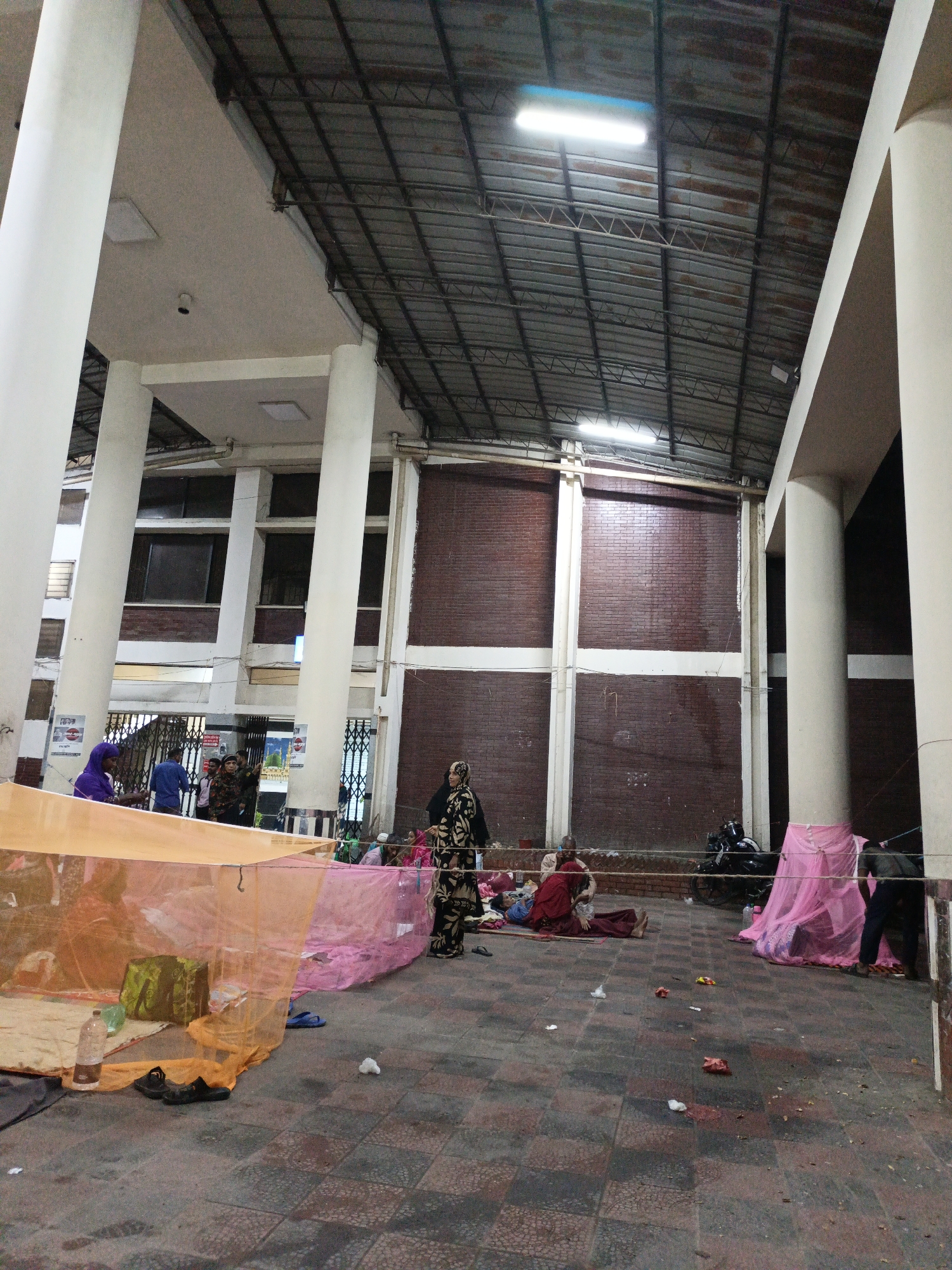 The image shows a large, open indoor space in Unknown, Unknown, likely a waiting area or public transportation hub. The architecture includes tall, white columns supporting a high, metal roof with visible support beams and fluorescent lighting. The walls are constructed of red brick in a segmented pattern. Several people are present, some lying on blankets or mats under mosquito nets of varying colors (orange and pink). A woman in a patterned black and gold garment stands among the people, while others are visible further in the background near a gated entrance. A motorcycle is parked against the brick wall. Debris, including plastic bottles and wrappers, litters the tiled floor. The overall impression is one of temporary shelter or a makeshift resting area in a public building.