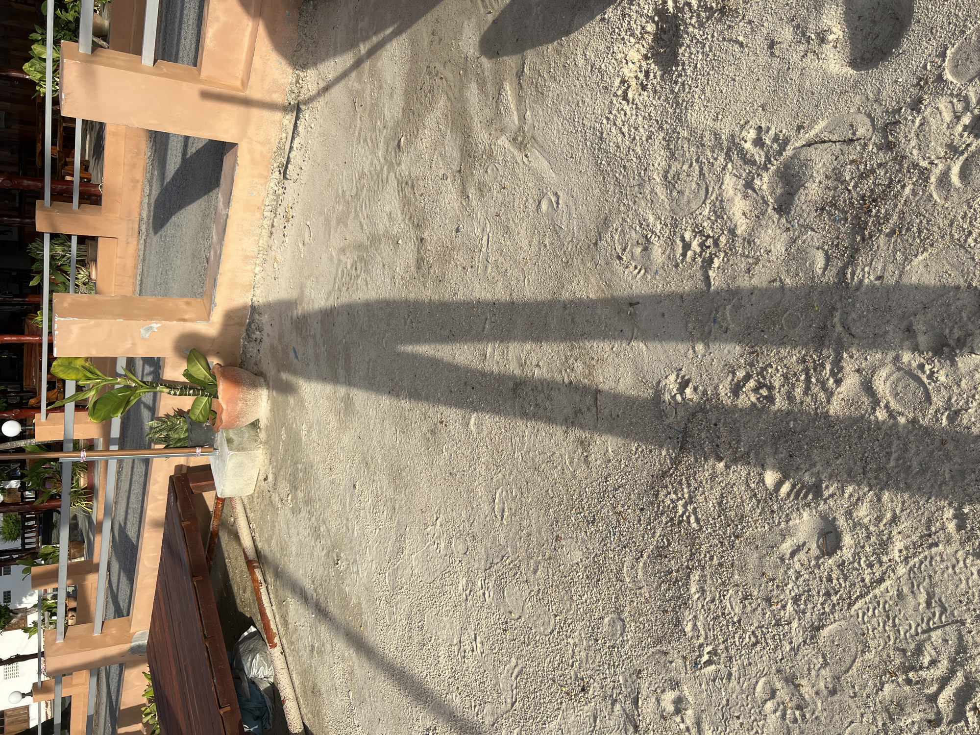 The image depicts a sandy ground with scattered footprints and debris. A long, distinct shadow of a person is cast across the sand, stretching from the bottom of the frame upwards. The shadow's outline suggests the person is standing and facing away from the light source. In the background, a concrete structure with a terracotta-colored railing and several potted plants are visible. A wooden platform is also present on the left side of the image. The lighting suggests it is either early morning or late afternoon, casting long shadows.