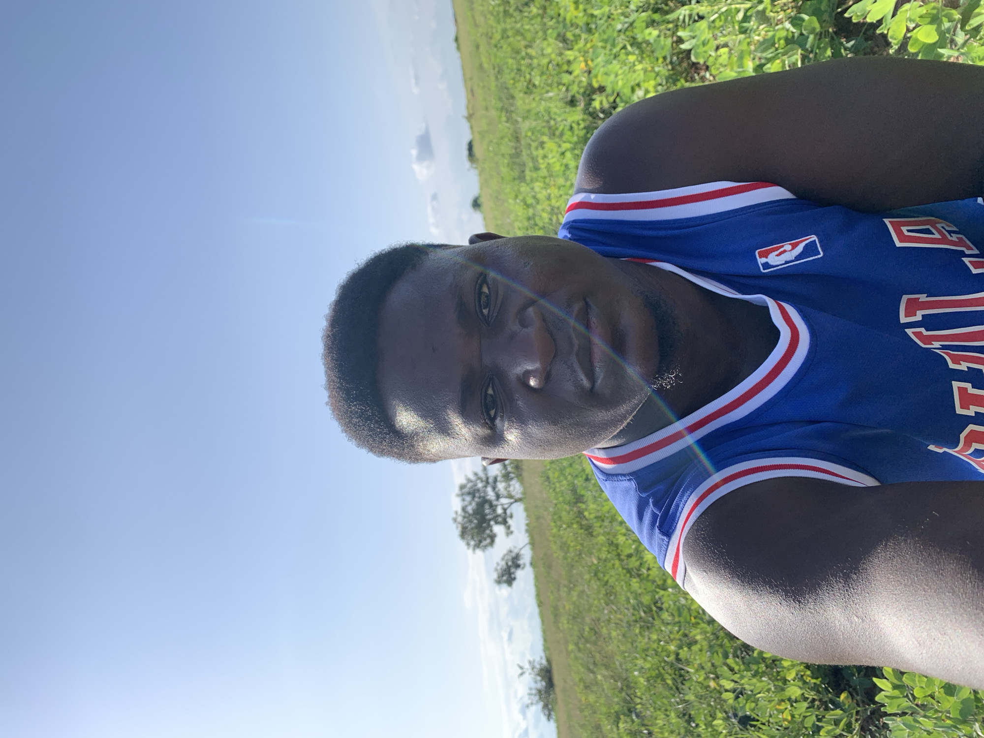 A man wearing a blue basketball jersey with red and white trim is pictured in a field of green plants under a clear sky. The man is looking directly at the camera. The background shows a slightly hilly landscape with a few trees in the distance. The sun is bright, casting shadows and creating a lens flare effect in the image. The location is the city of Akwanga, Nigeria.