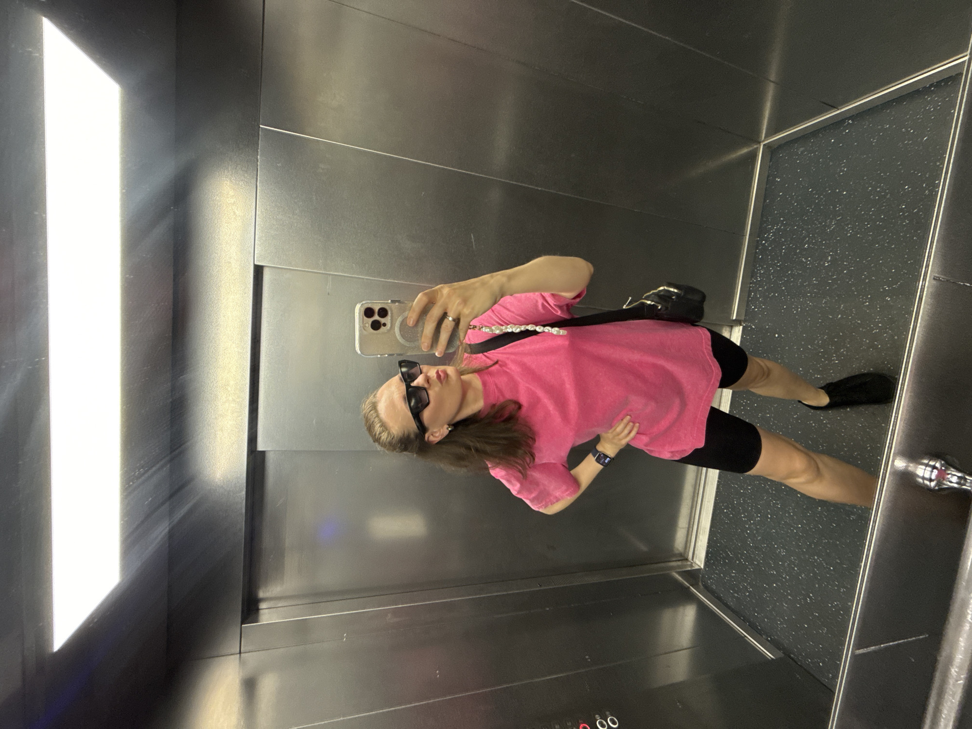 An adult female with brown hair pulled back is standing in an elevator, taking a selfie. She is wearing a bright pink oversized t-shirt, black biker shorts, black sneakers, and black sunglasses. A black purse with a pearl strap hangs over her shoulder. Her left hand is on her hip. The elevator has metallic walls and a speckled dark grey floor. The lighting fixture on the ceiling is a bright rectangular panel. The image does not provide any explicit visual cues to suggest the location is Saint Petersburg, Russia.