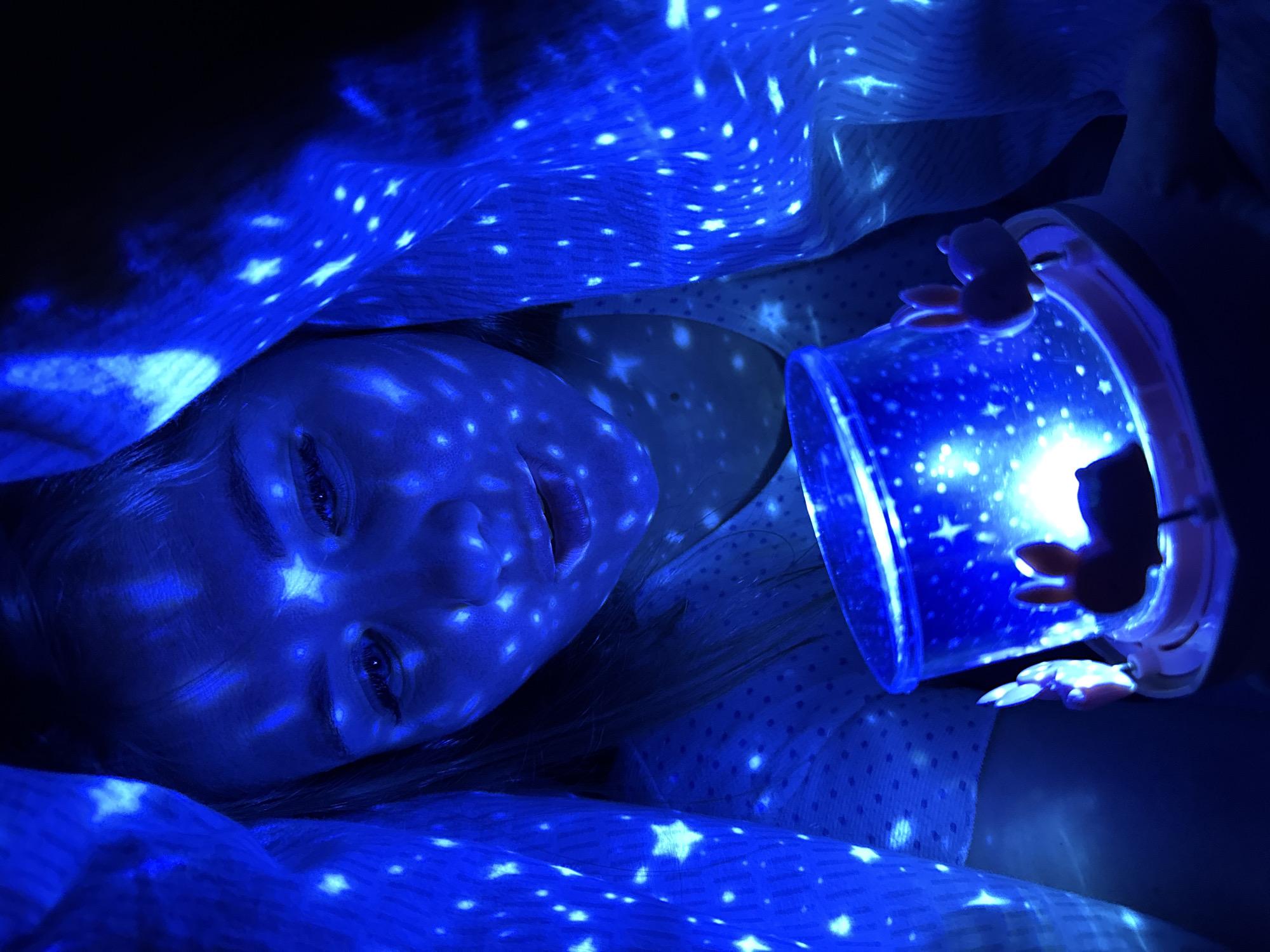 This image, captured in Gornyi (Dvurechenskoe S/P), Russia, displays a low-light indoor scene dominated by blue illumination. A person, likely a young individual with light hair, is positioned centrally, their face and upper torso illuminated by numerous projected blue star and circular light patterns. Their gaze is directed downwards, mouth slightly agape, towards a cylindrical light projector held in their hands. This device, featuring small, stylized bunny figures around its upper rim, emits a bright blue glow and projects the aforementioned star and dot patterns onto the surrounding surfaces. A light-colored, textured fabric, possibly a blanket or sheet, is draped over the individual and extends into the foreground and background, also displaying the projected blue light patterns. The individual wears a light-colored garment with faint polka-dot patterns. The background is indistinctly dark, emphasizing the projected light effects.