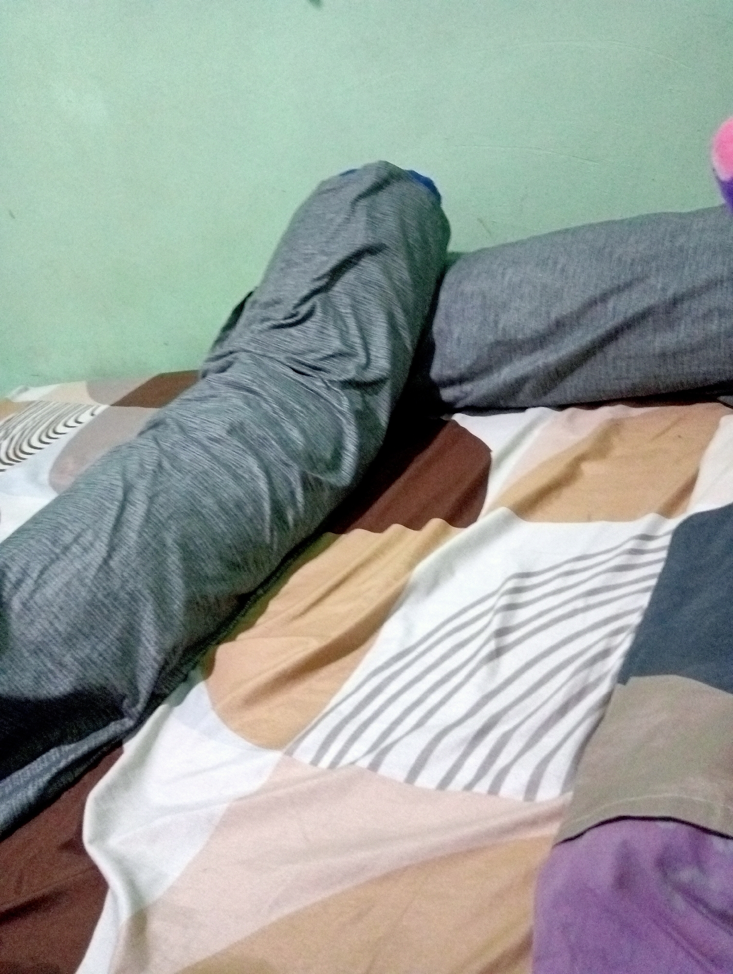 The image depicts a close-up view of a bed with patterned sheets and two cylindrical pillows. The pillow on the left is covered in a grey fabric with a subtle striped pattern, and it appears to be positioned horizontally across the bed. To its right, another grey pillow is visible, also cylindrical and positioned horizontally. The bedsheets have a diagonal pattern featuring blocks of white, beige, brown, and grey stripes. In the upper right corner of the image, a small portion of a pink object is visible, possibly a stuffed animal or part of another pillow. The background wall is painted a light green color. The image does not provide any specific information that would allow for identification of its location within Kota Negara Timur, Indonesia.