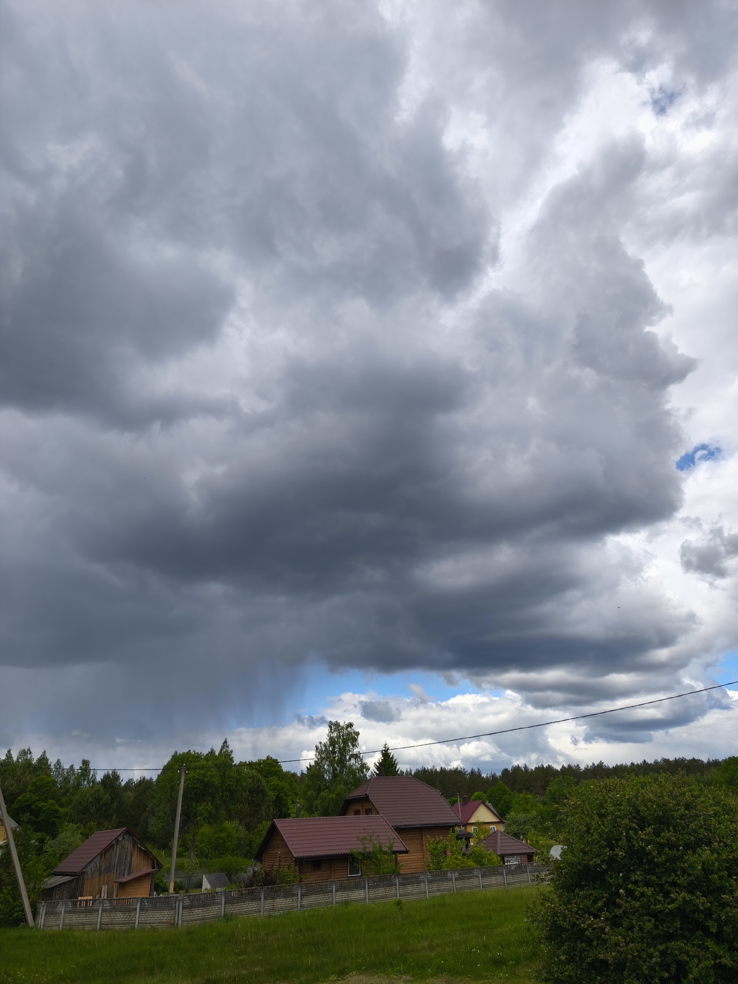 The image shows a rural scene in Парэчча, Belarus. Overcast skies filled with dark, ominous storm clouds dominate the view. A brief shower appears to be falling in the distance beneath the clouds. The landscape features wooden houses with reddish-brown roofs surrounded by green vegetation and a concrete fence. A forest lines the horizon. Power lines stretch across the sky.