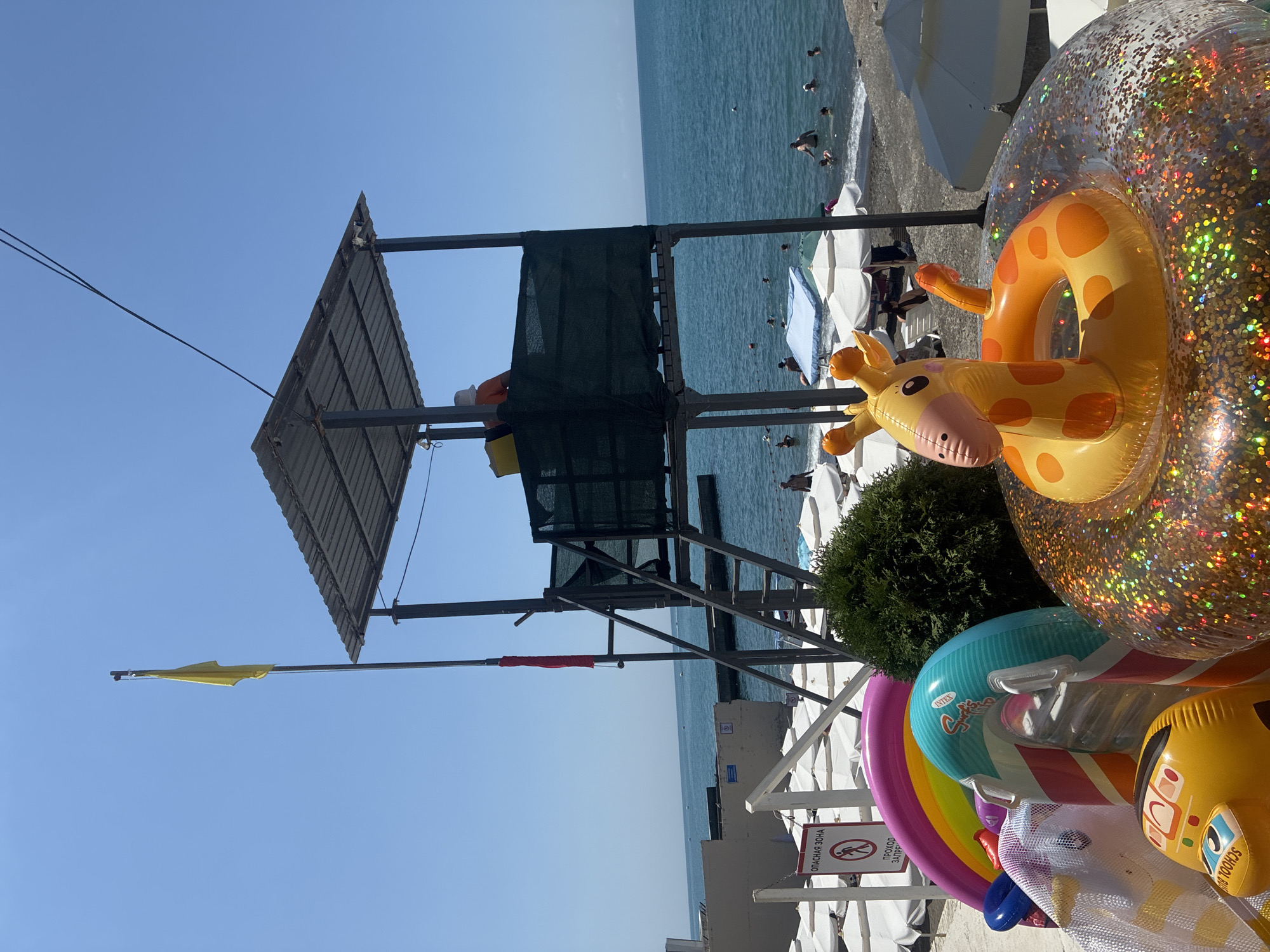 The image depicts a crowded beach in Sochi, Russia, on a clear, sunny day. In the foreground, a collection of colorful inflatable toys, including a rainbow-shaped float, a unicorn, a yellow school bus, and a giraffe-shaped float, are piled up. Behind the inflatables, a lifeguard tower stands tall, with a yellow flag flying from its top. A lifeguard, wearing an orange shirt and a white hat, is visible in the tower. The beach is lined with white lounge chairs and umbrellas, and the blue sea stretches out to the horizon, dotted with swimmers. The overall scene is one of a typical summer day at a popular beach resort.