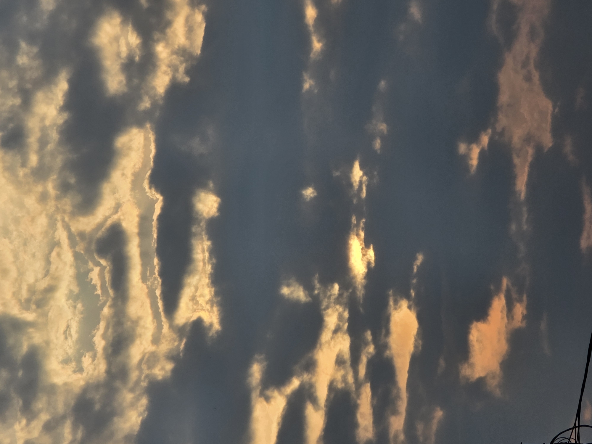 The image depicts a cloudy sky at dusk, with the sun partially visible, casting a warm, golden glow through breaks in the dark, ominous clouds. The scene appears to be an outdoor, natural setting with no people or specific man-made objects clearly identifiable in the frame, except for a partial view of what seems to be electrical wires or cables in the lower left corner, suggesting an urban or semi-urban environment. The overall impression is one of dramatic natural beauty, possibly indicating an approaching storm or the end of a day. The metadata for the image does not specify a city, but if this image is indeed from Ермолаево, Russia, it captures a typical atmospheric condition for that region during the evening hours.