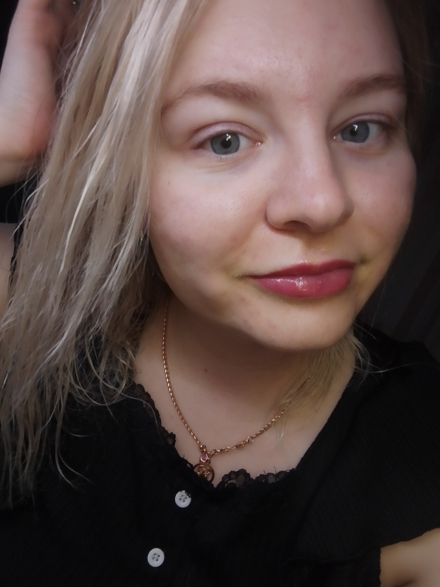 A young woman with blonde hair and blue eyes is captured in a close-up selfie. She wears a black top with lace detailing at the neckline and two visible white buttons. A delicate gold necklace with a circular pendant adorns her neck. Her lips are glossy with a pink-toned lip product. She is likely indoors, but the location in Izhevsk, Russia is not discernible from the visual information available.