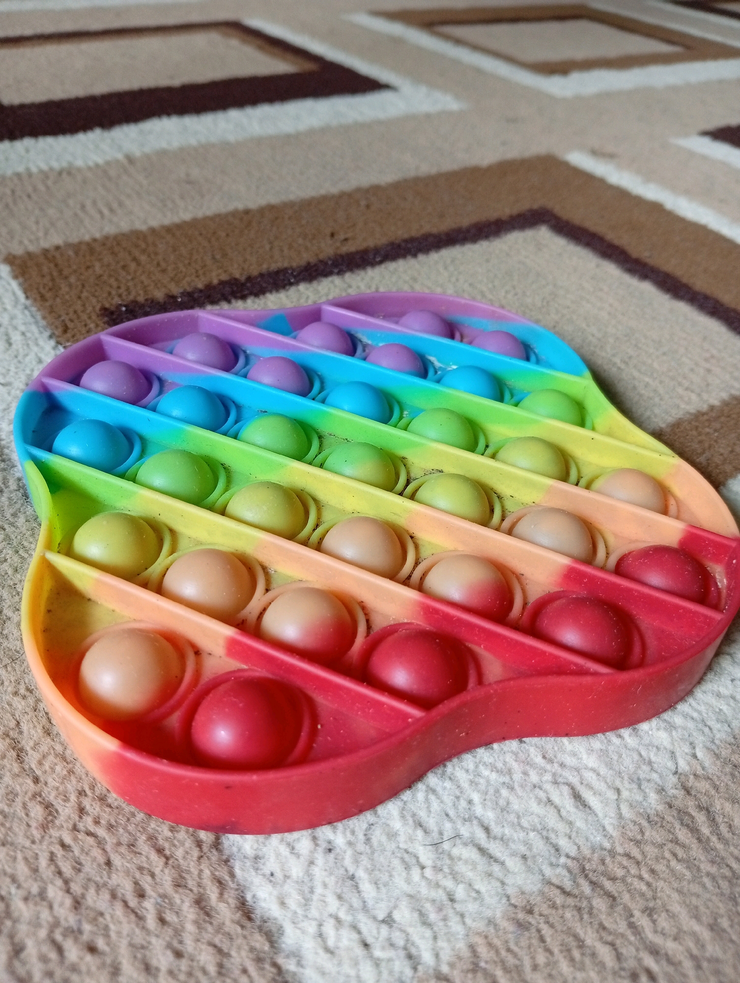 An overhead shot shows a colorful pop-it toy shaped like a heart, made of silicone and filled with small, round bubbles. The toy is positioned on a carpet with a geometric pattern of brown, beige, and white stripes. The pop-it toy displays a gradient of colors from purple and blue at the top to green, yellow, orange, and red at the bottom. Each bubble appears to be a different shade within these color bands. The toy lies flat on the carpet, with the lighting casting soft shadows on the bubbles. The background is out of focus, emphasizing the toy as the main subject. There is no indication of the city of Kebondalem, Indonesia in the image.