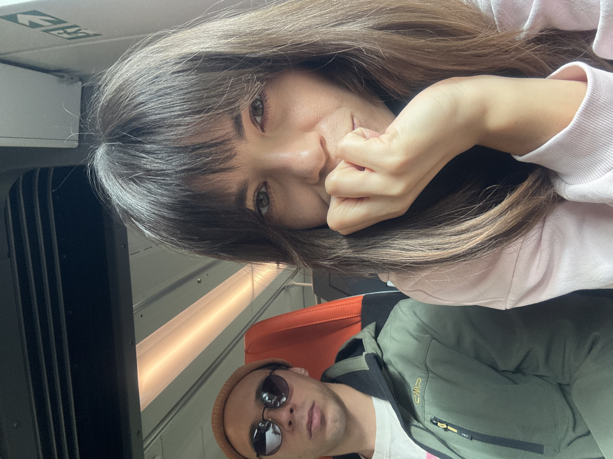 A medium shot captures two people sitting next to each other on a train, with the right side of the frame focusing on a woman's face as she rests her hand on her chin. She has long, dark brown hair with bangs, and is wearing a light pink hooded sweatshirt. To her left, a man with short brown hair is wearing a tan beanie, sunglasses, and a green jacket over a white t-shirt. The background shows the interior of a train car, with orange seats visible and a long light fixture running overhead.