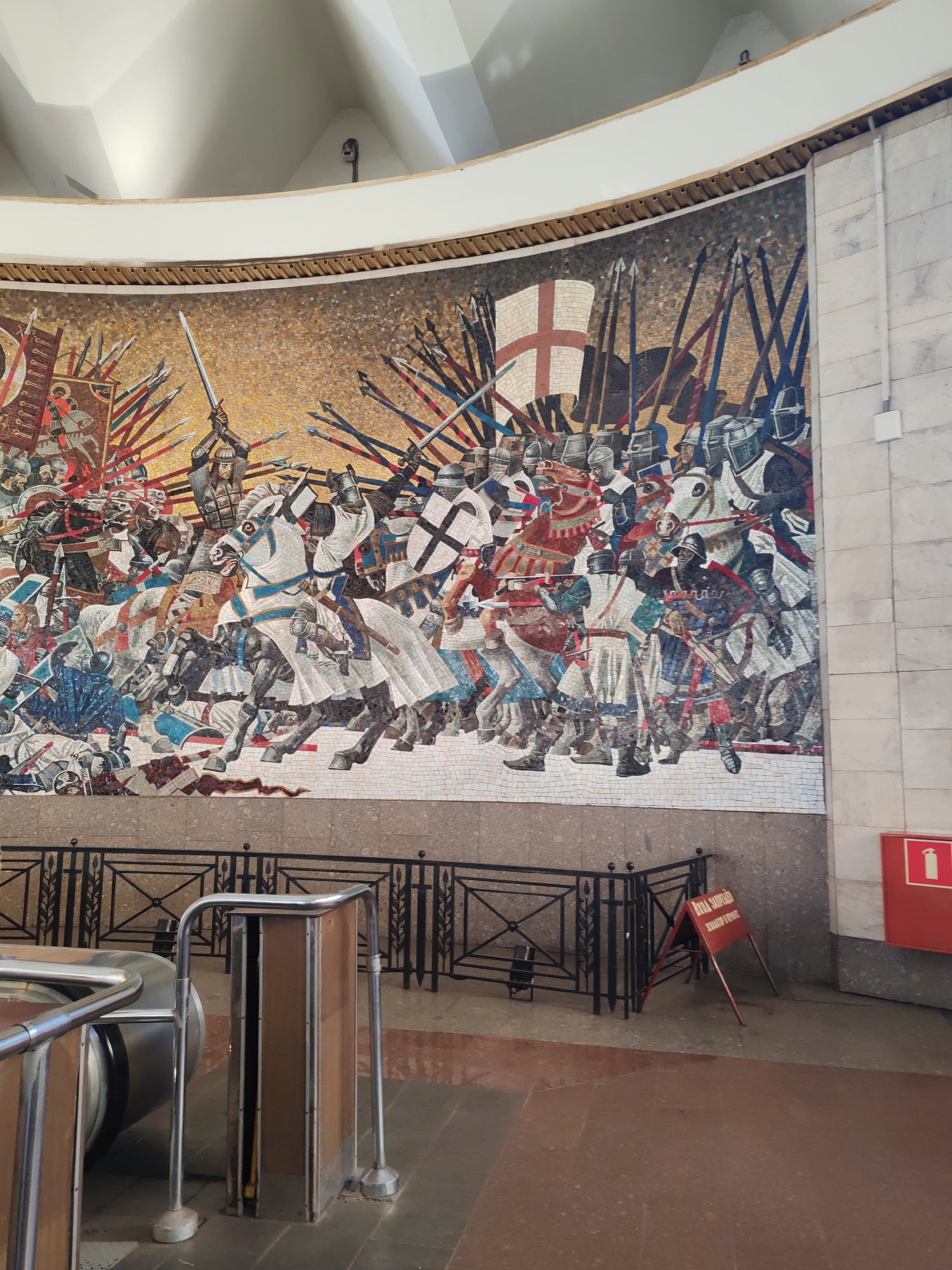 The image displays a large, vibrant mosaic mural depicting a fierce medieval battle scene, set into a curved wall of a grand indoor space. The mosaic dominates the upper half of the frame, showcasing two armies clashing with dynamic energy. Figures are rendered on horseback and on foot, clad in various forms of armor and helmets, wielding swords, spears, and axes. A prominent central figure on a white horse is depicted raising a sword. On the right side of the mosaic, a large white flag bearing a red St. George's Cross stands amidst a dense array of weaponry. Another banner with a red and gold design is visible on the left. The mosaic employs a rich color palette of blues, reds, whites, and browns, set against a golden and brown background.
Below the mosaic, a black decorative metal railing runs along its base. The floor in the foreground is tiled in a reddish-brown hue, transitioning to a lighter beige or gray towards the right wall. In the bottom left, part of a modern silver metal railing structure, possibly associated with an escalator or barrier, is visible. The right side of the image reveals a light-colored, possibly marble or stone, tiled wall extending upwards. The ceiling above the mosaic is high, curved, and white.
A red sign indicating a fire extinguisher is mounted on the tiled wall to the right. On the floor, a red A-frame sign is visible, bearing the text "ВХОД ЗАПРЕЩЁН" (Entry Forbidden) and "ОПАСНО ДЛЯ ЖИЗНИ" (Dangerous to Life). The setting suggests a public building or metro station in Saint Petersburg, Russia, given the scale and artistic style.
