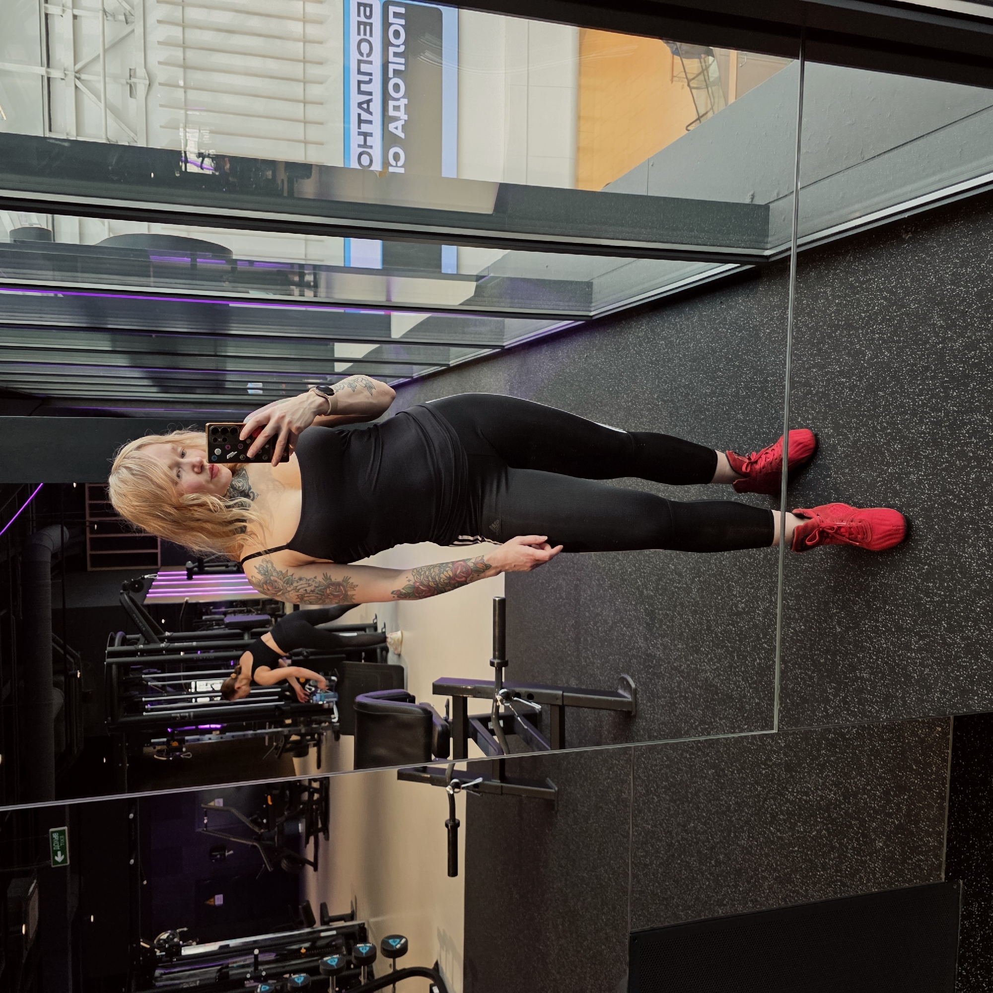 A young woman wearing athletic apparel and red sneakers is taking a selfie in a mirrored wall at a gym. Behind her is a variety of gym equipment, and in the reflection, the exterior view includes buildings with signs in Russian. Given the text in the sign, the location is likely in Saint Petersburg, Russia. Another person is working out in the background. The gym has black flooring and purple lighting accents.