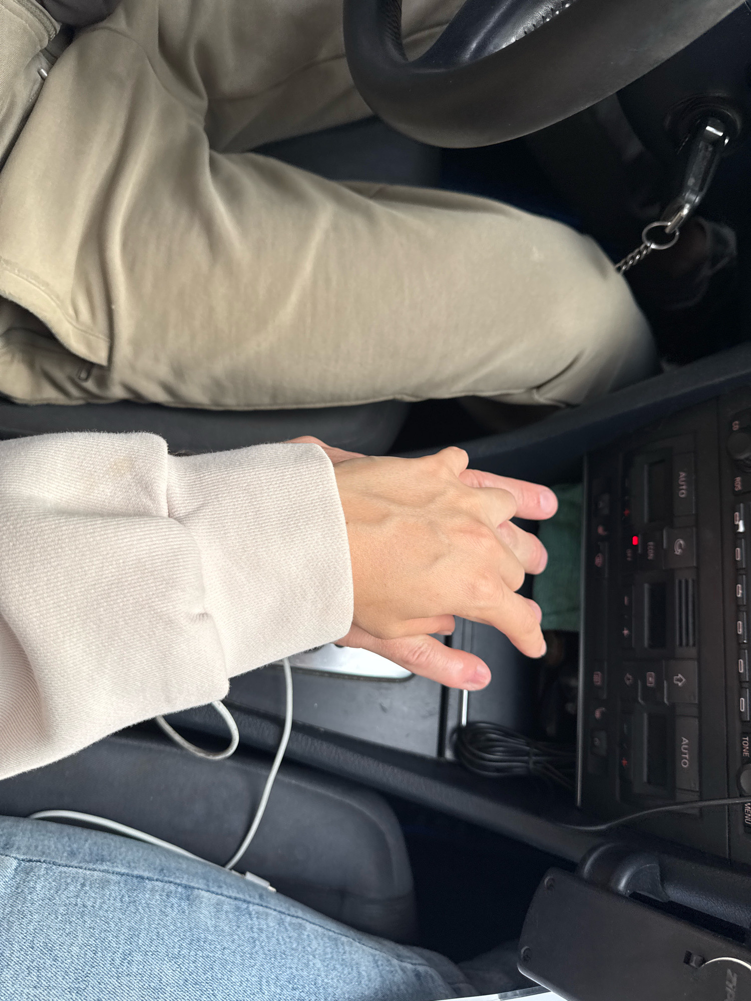 A top-down view shows the hands of two individuals interlocked in the center console of a car. One person, wearing a light-colored hoodie, has their left hand resting on the gear shift, with fingers interlaced with the other person's right hand. The second person, wearing tan pants, has their right hand on top of the first person's left hand. The car's dashboard, including the gear shift, buttons, and a charging cable, is visible. The scene appears to be within a vehicle, possibly in the city of Homyel, Belarus.