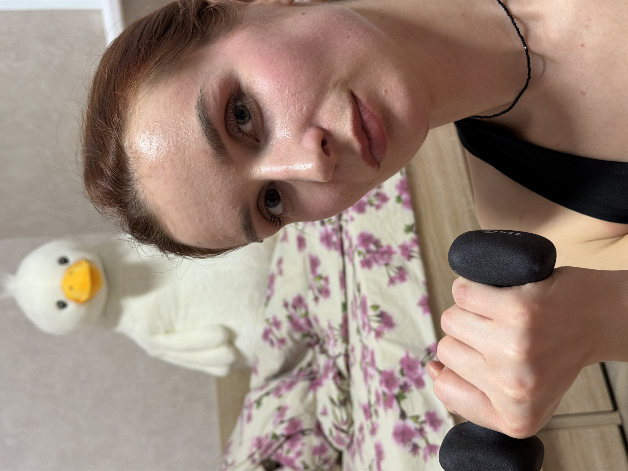 A woman with auburn hair and fair skin holds a dumbbell, looking directly at the camera. She wears a black necklace and a black sports bra. The background includes a floral-patterned garment, a white plush duck toy with a yellow beak, and what appears to be a wooden surface. No information is given in the image that relates to the location in the city of Chaikovsky, Russia.