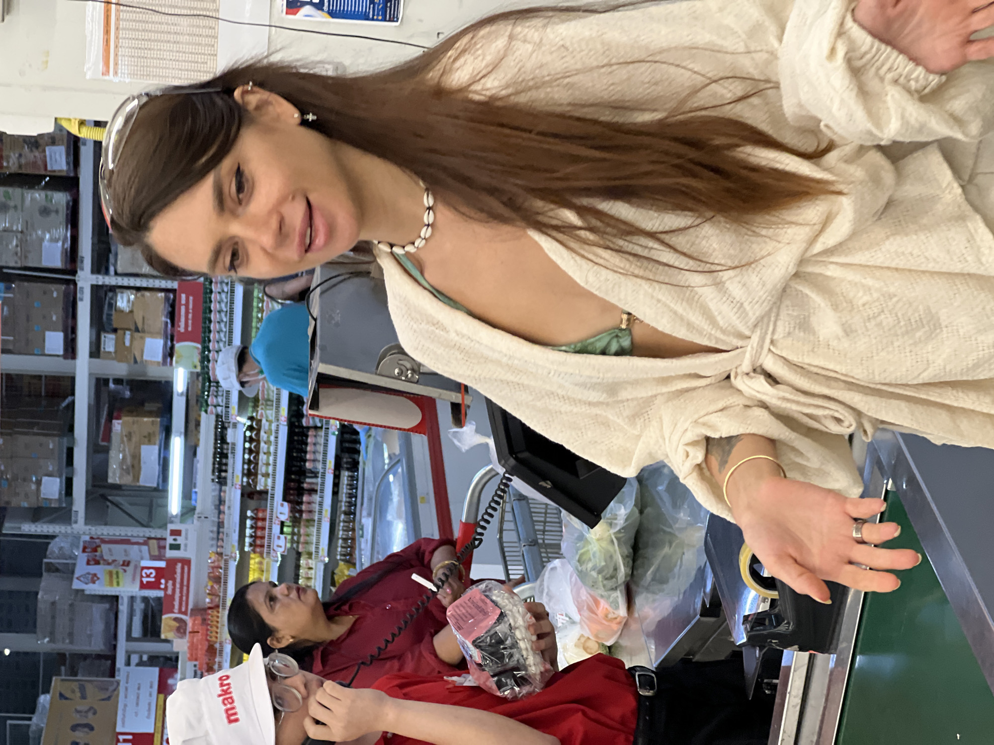 A woman with long brown hair, wearing a cream-colored open cardigan over a green bikini top and a shell necklace, stands at a checkout counter in a Makro store in Baan Bo Phut, Thailand. She has a hand extended, palm open, towards the camera, wearing a silver ring on her right index finger and a gold bangle on her left wrist. A cashier in a red shirt with a "makro" logo on their hat is to the left, holding a plastic-wrapped item. Behind them, another woman in a dark red shirt is visible. The checkout counter has a conveyor belt and a scanner. Shelves stocked with products are visible in the background.