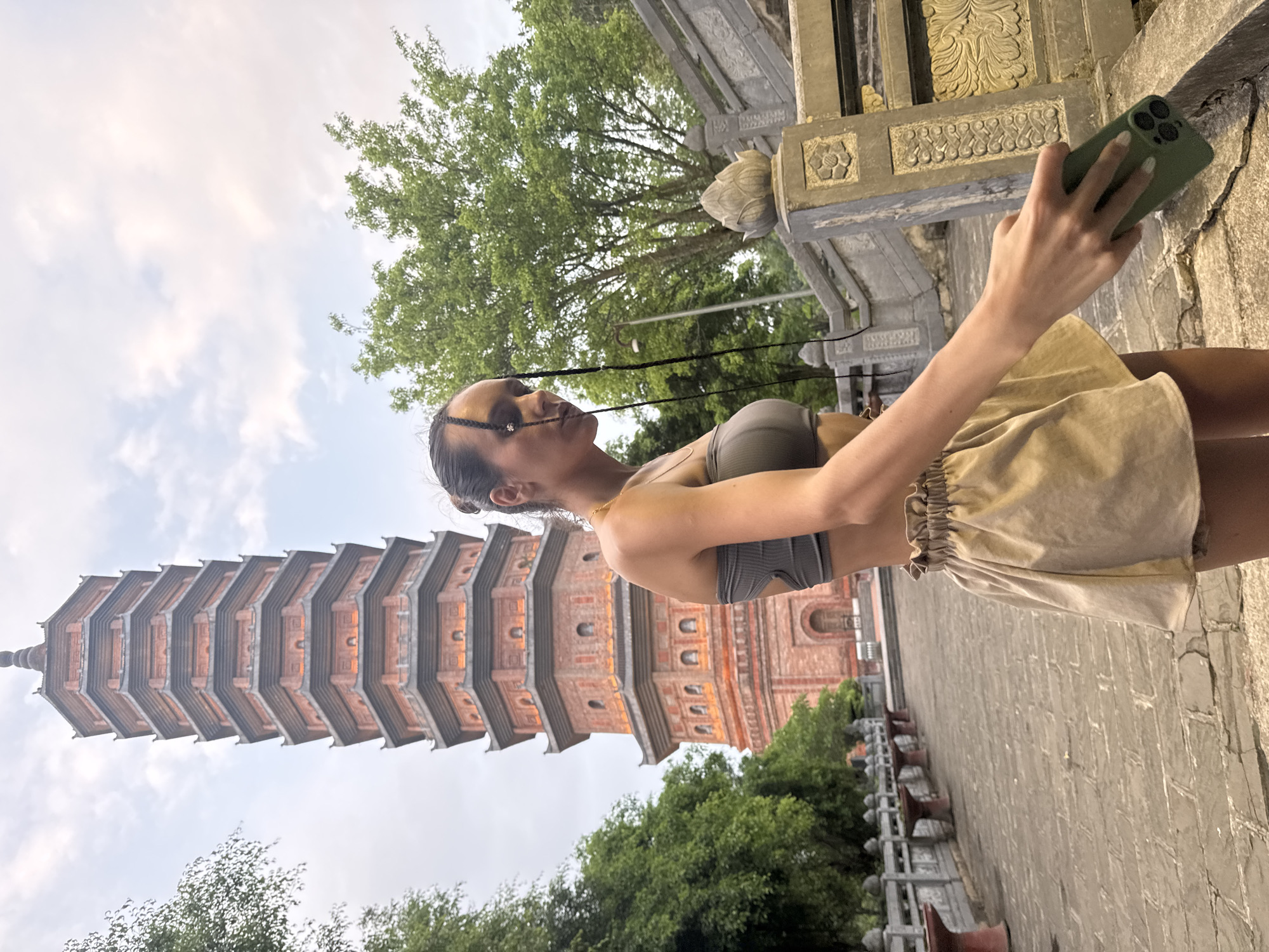 An image captures a woman standing on a stone pathway in front of a towering, multi-tiered pagoda. The pagoda, constructed of red brick, features several levels with decorative eaves and arched openings. Trees with lush green foliage surround the pagoda, and the sky above is partly cloudy. The woman is seen from the waist up, wearing a gray sports bra and a light brown skirt. Her hair is styled in braids that hang down her back. She is looking to her right and holding a green smartphone in her right hand. The pathway is paved with large stones, and to the right of the woman, there is a stone railing with decorative carvings. In the background, to the left, a row of stone benches is visible along the pathway. The setting appears to be a temple or historical site in Tây Hoa Lư Ward, Vietnam, likely a tourist destination.
