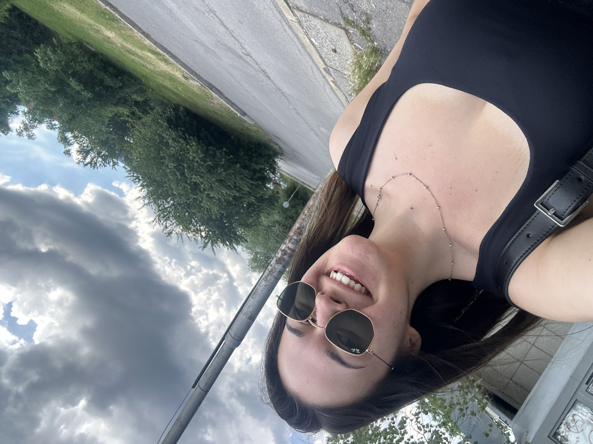 The image is a selfie taken outdoors, likely in Sofia, Bulgaria, given the context. A young woman with long dark hair and sunglasses is smiling at the camera. She is wearing a black tank top and a delicate silver necklace. The background shows a cloudy sky, trees, a grassy slope, and a paved road. A metal pole is visible on the left side of the frame. The scene appears to be a casual, everyday moment.