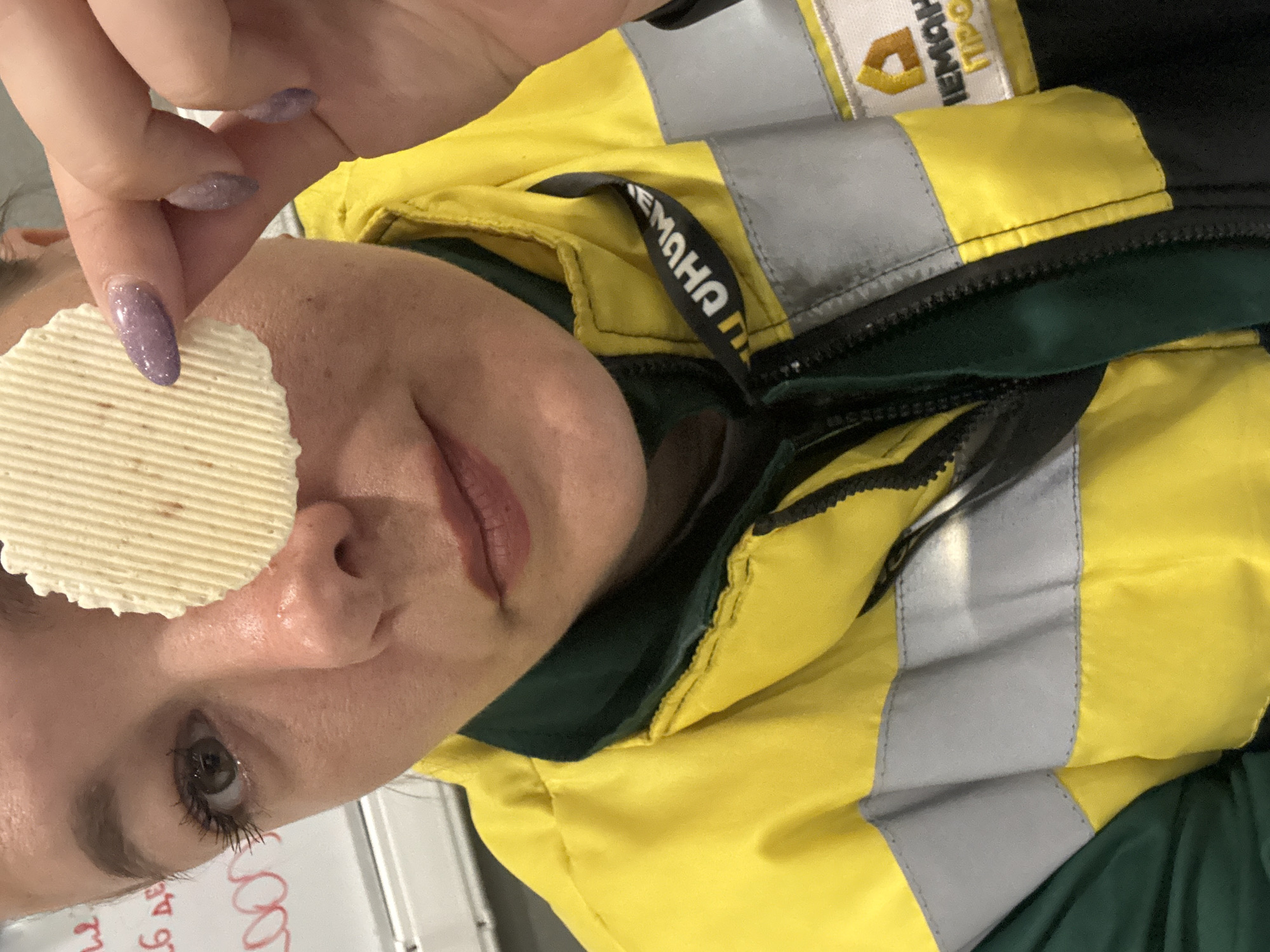 A woman is holding a ridged potato chip over her left eye. She has light skin, brown eyes, and lavender nail polish. She is wearing a yellow and grey high-visibility jacket with black and green accents. The jacket has Russian writing on the collar. The location cannot be confirmed as the city of Lupanovo, Russia.
